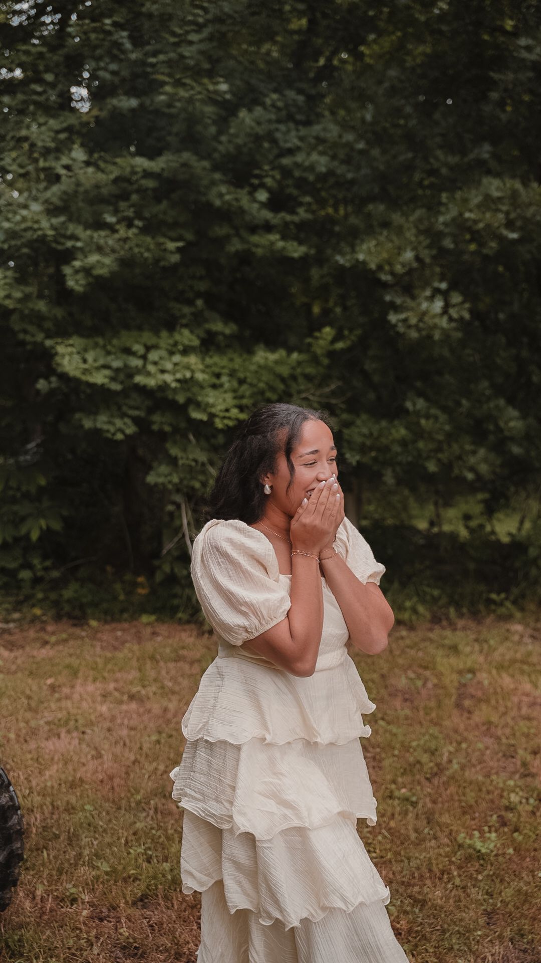 Young woman in white ruffled dress standing in a field, covering her mouth with both hands, with green trees behind her.