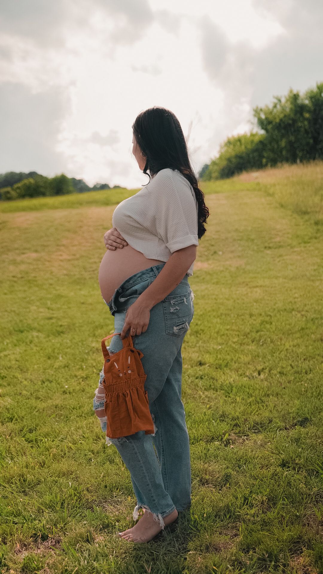 Pregnant woman in jeans and white top holds baby shoes while standing barefoot in a green field at sunset.