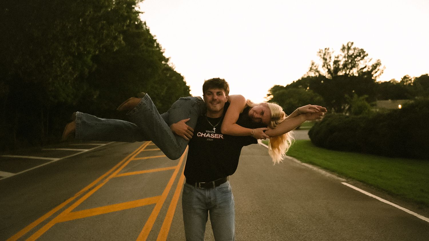 A young man lifts a blonde woman in his arms on a tree-lined road at sunset.