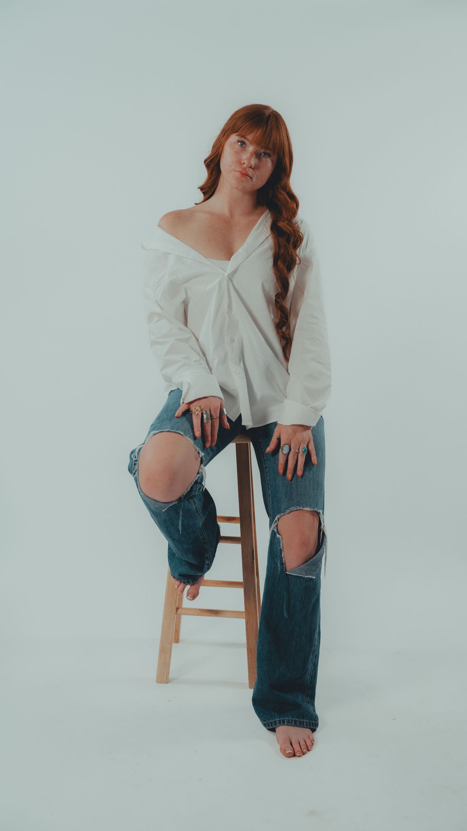 A redhead woman poses confidently in a white off-shoulder shirt and ripped jeans, leaning against a wooden stool.