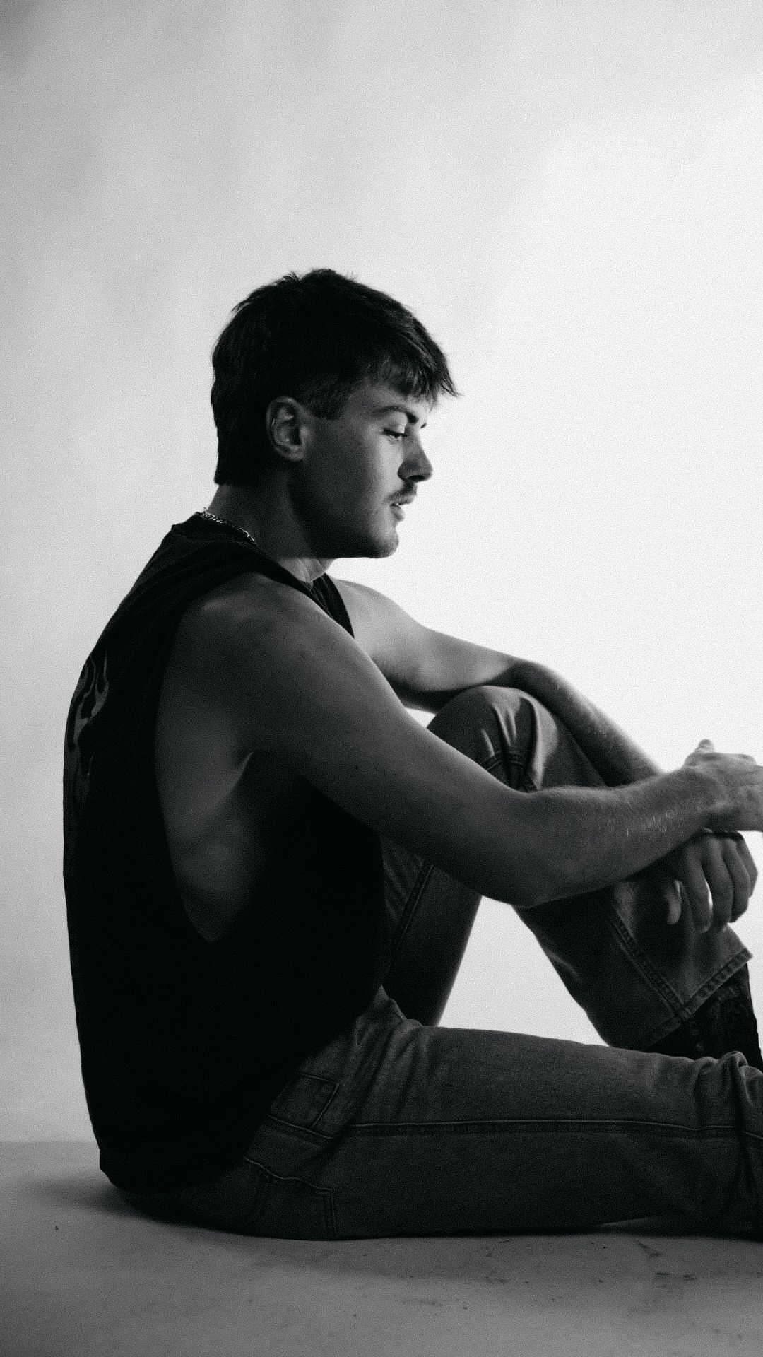 Black and white photo of a young man in a tank top and jeans sitting on the floor with knees up, looking down.