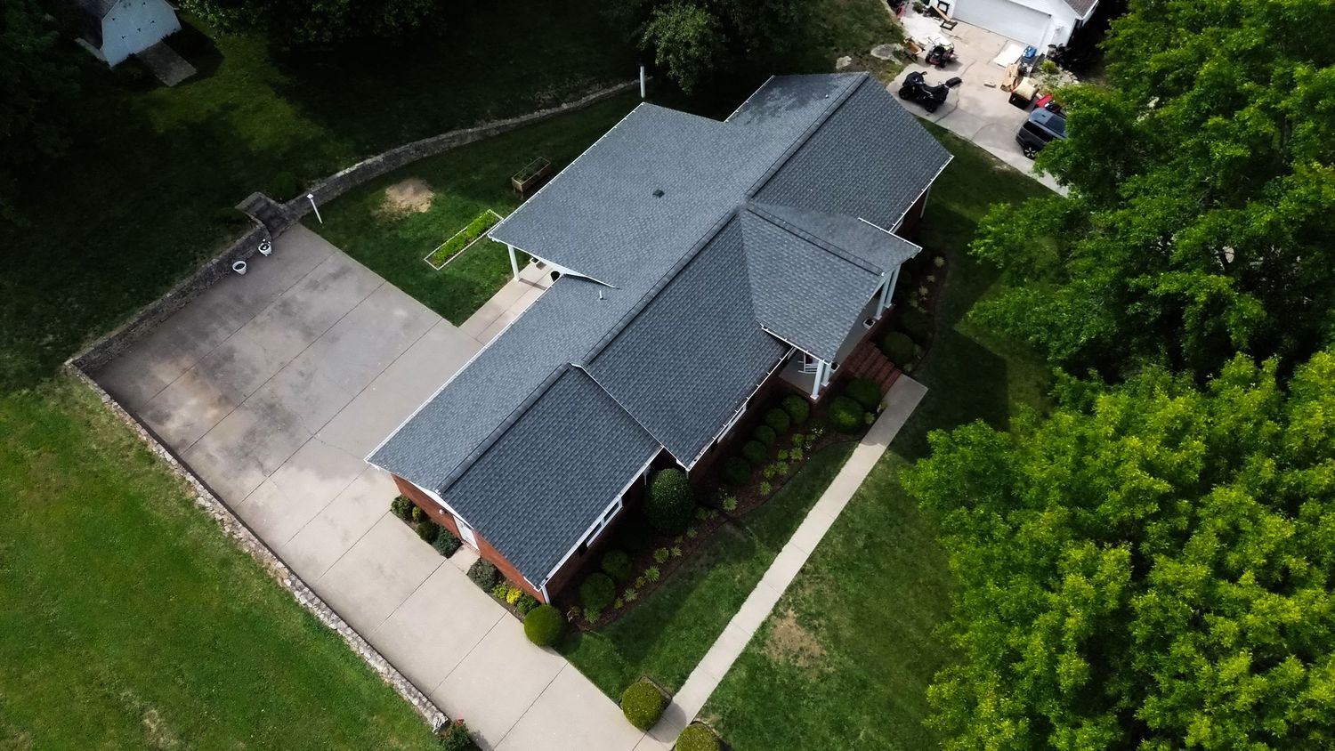Aerial drone view of a single-story suburban home with gray roof, concrete driveway, and lush green trees surrounding the property.