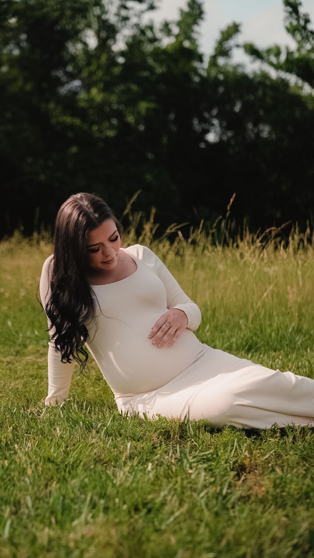 Pregnant woman in elegant white long-sleeve dress sitting on green grass, cradling her baby bump.