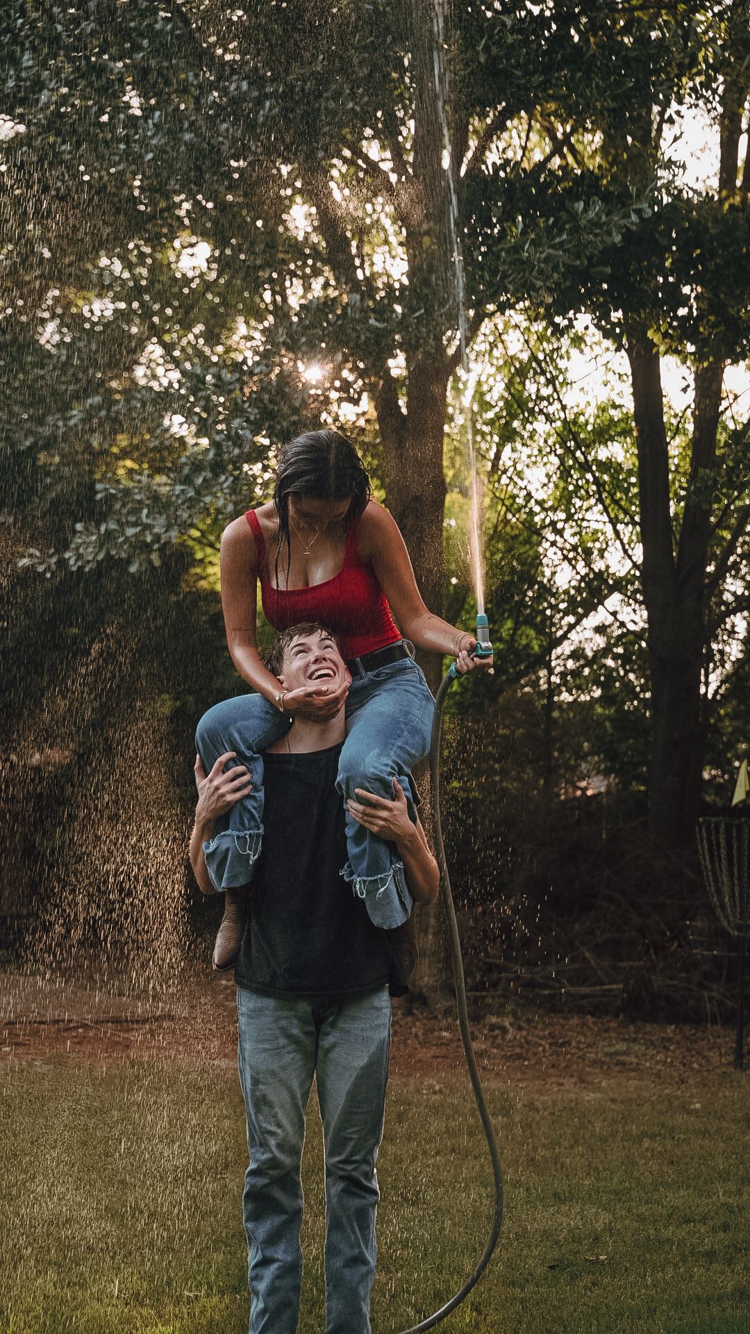 A laughing couple plays with a garden hose outdoors, woman sitting on mans shoulders, surrounded by lush green trees at sunset.
