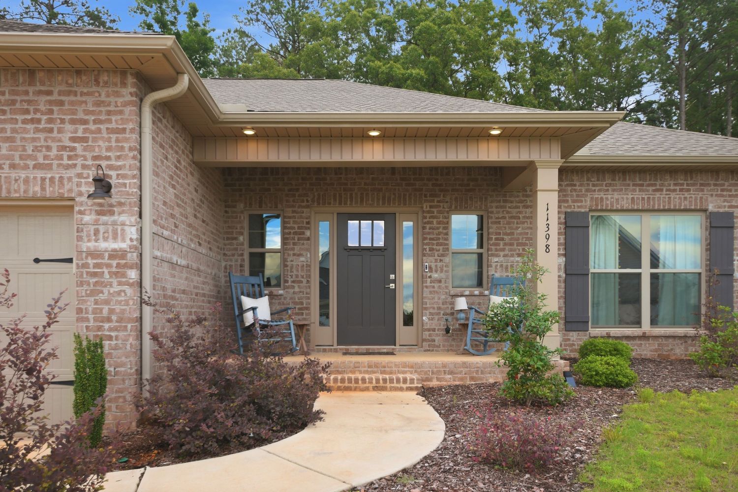 Brick ranch home exterior with covered front porch, dark front door, and lush landscaping surrounded by mature trees.
