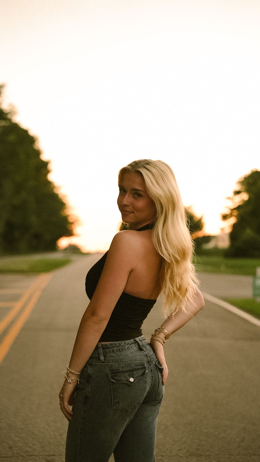 Blonde woman in black tube top and jeans poses on a road at golden hour sunset with trees in background.