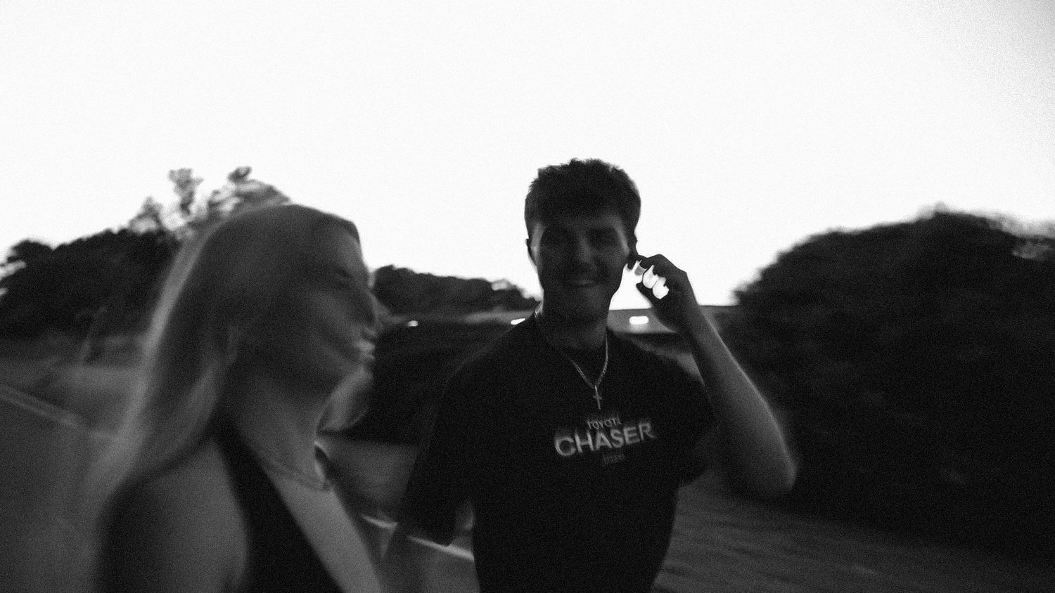 Black and white photo of a young man in a Chaser t-shirt and a blonde woman outdoors at dusk.