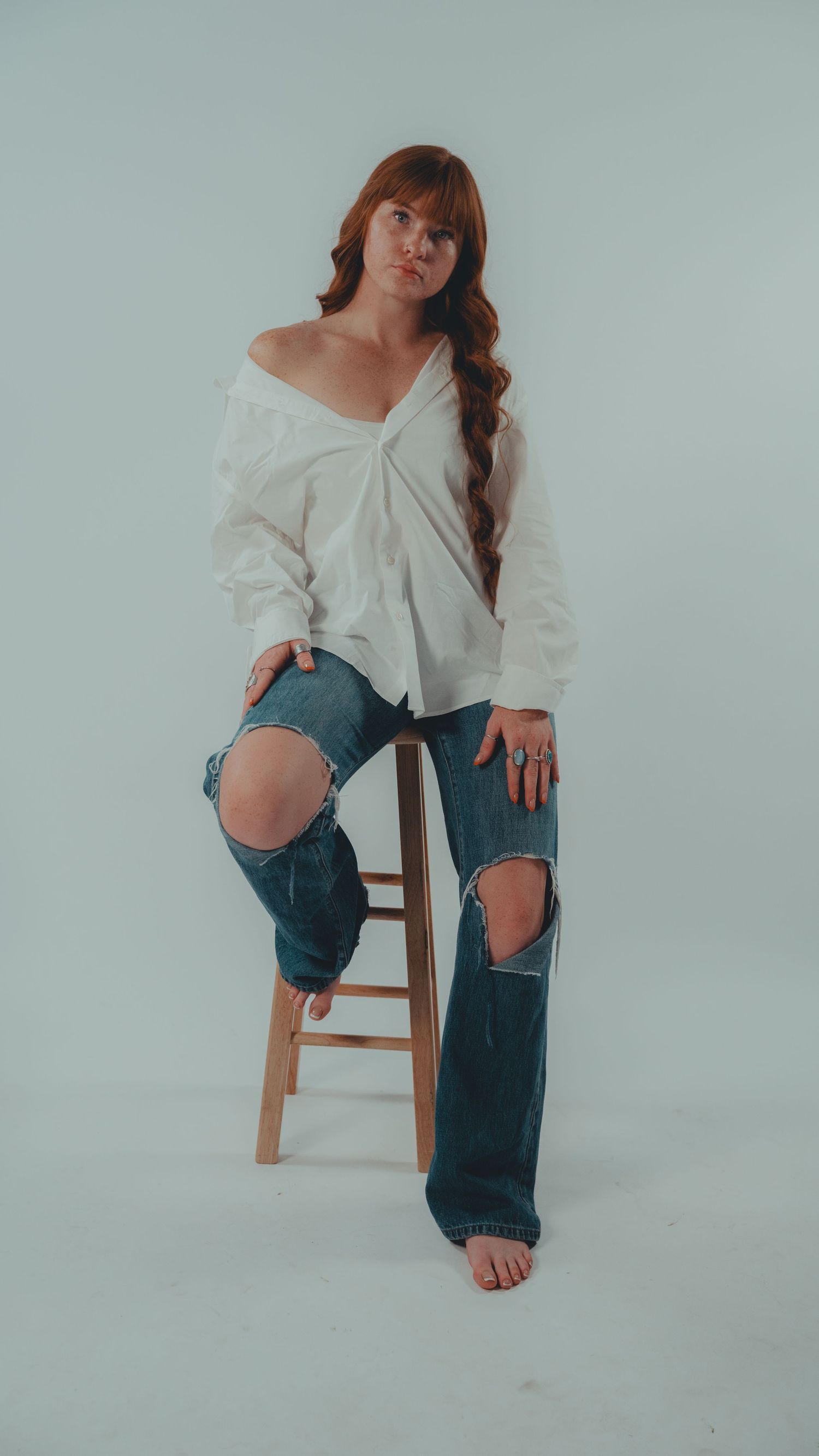 Woman with red hair in white off-shoulder top and ripped jeans poses on wooden stool against light gray background.