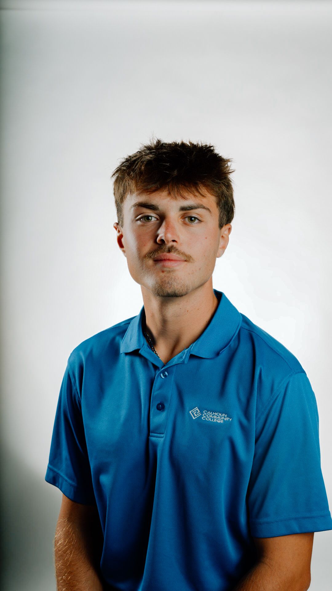 Young male student in blue Calhoun Community College polo shirt posing for portrait against white background.
