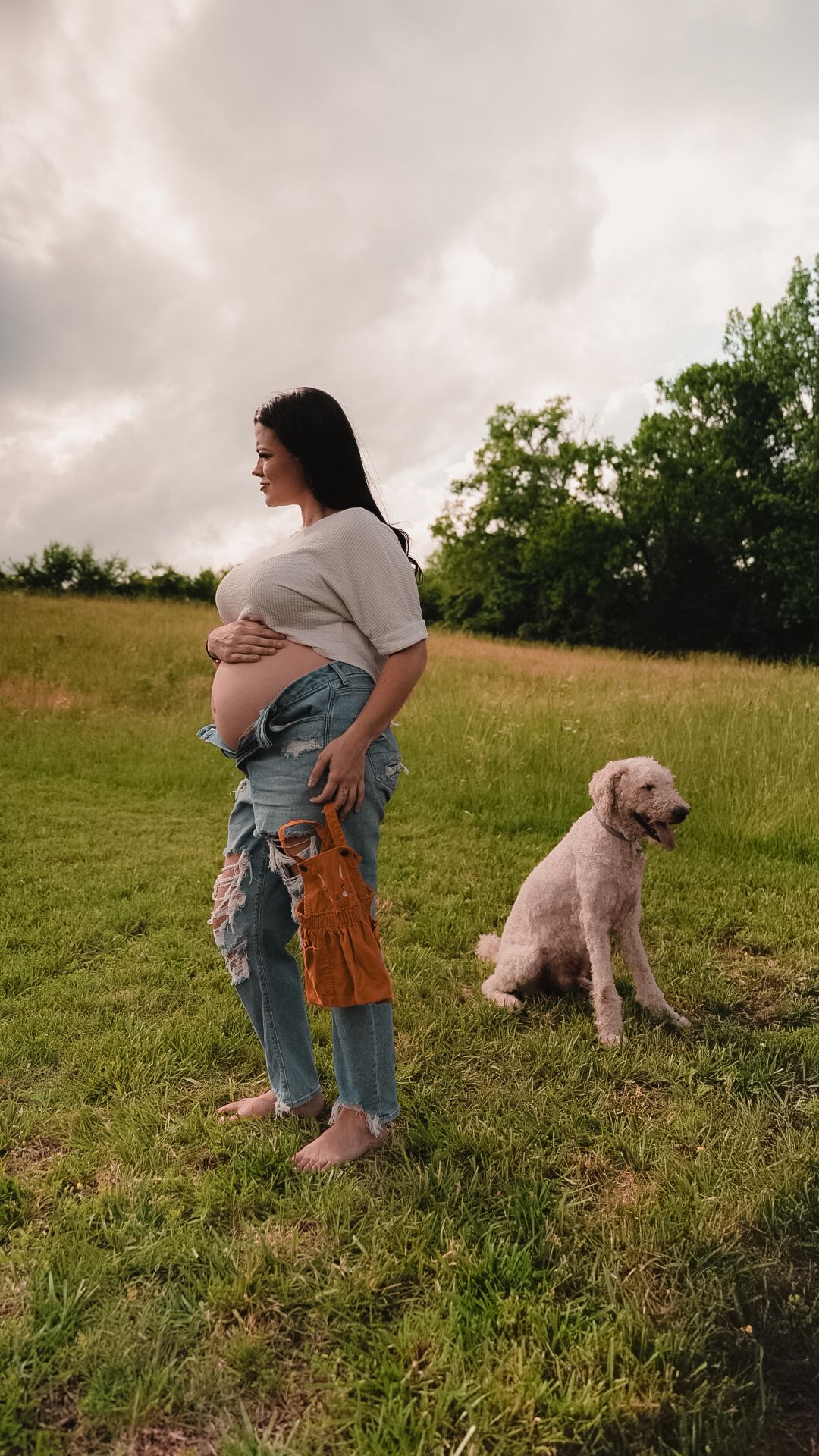Pregnant woman in jeans holds belly outdoors on green field with fluffy dog beside her under cloudy sky.
