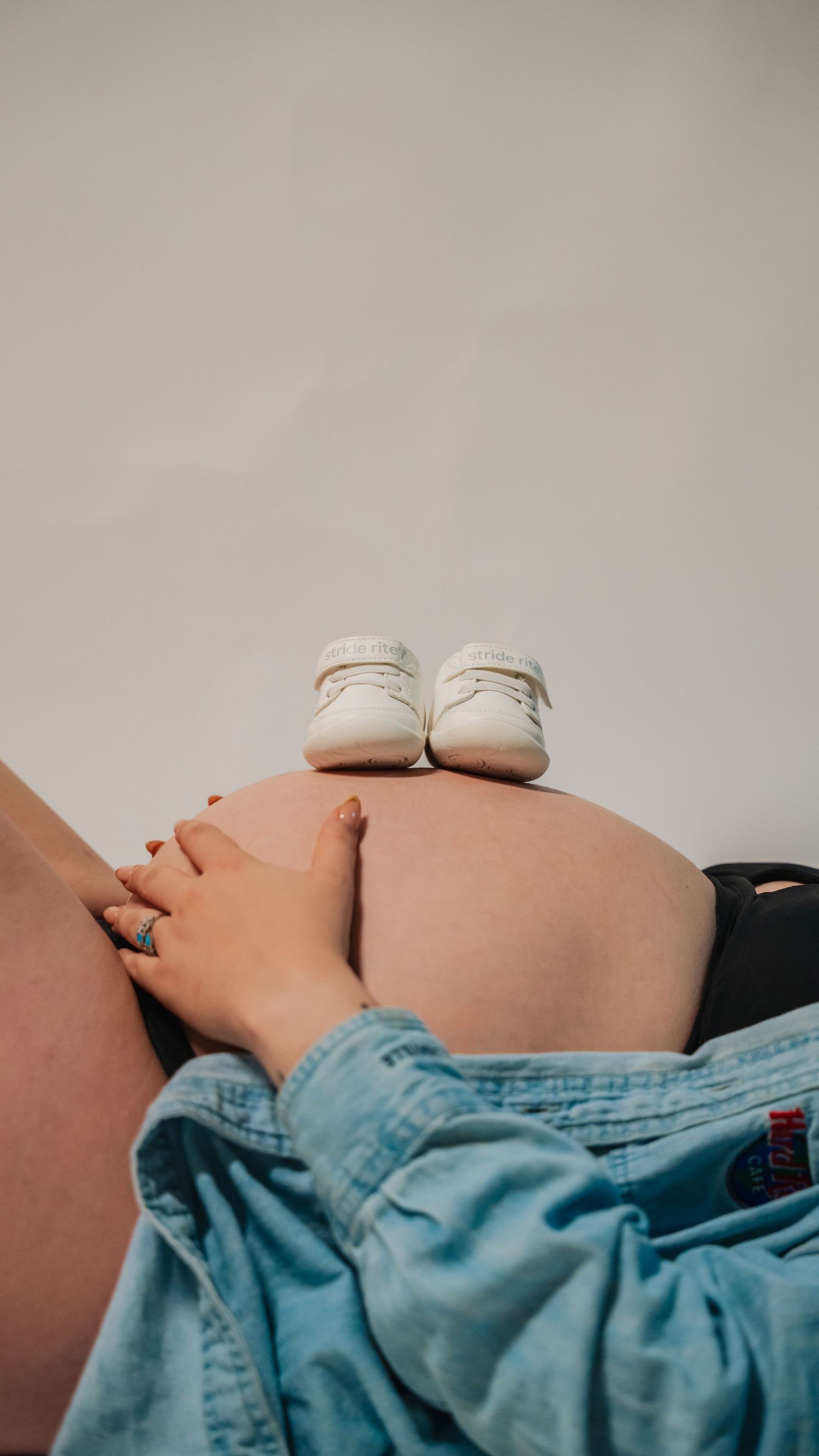 Pregnant woman resting with tiny white baby shoes balanced on her bare baby bump, wearing denim jeans.