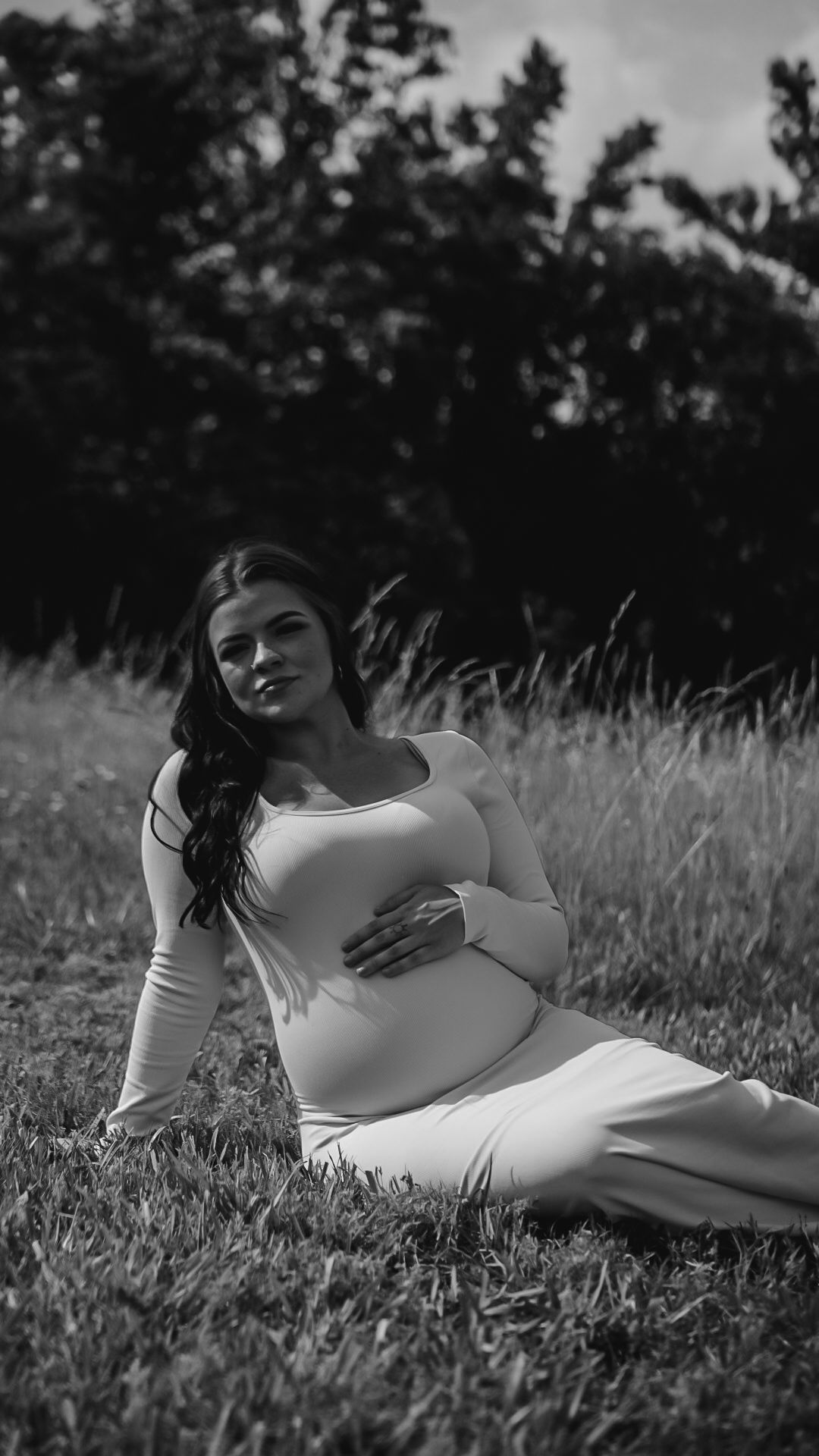 Black and white photo of pregnant woman in white dress sitting on grass, holding her baby bump.