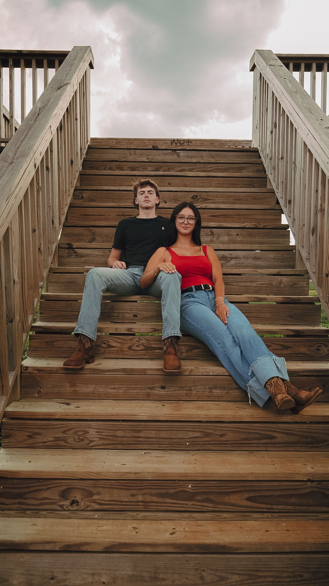A young couple sits together on wooden stairs outdoors, wearing jeans and boots under a cloudy sky.