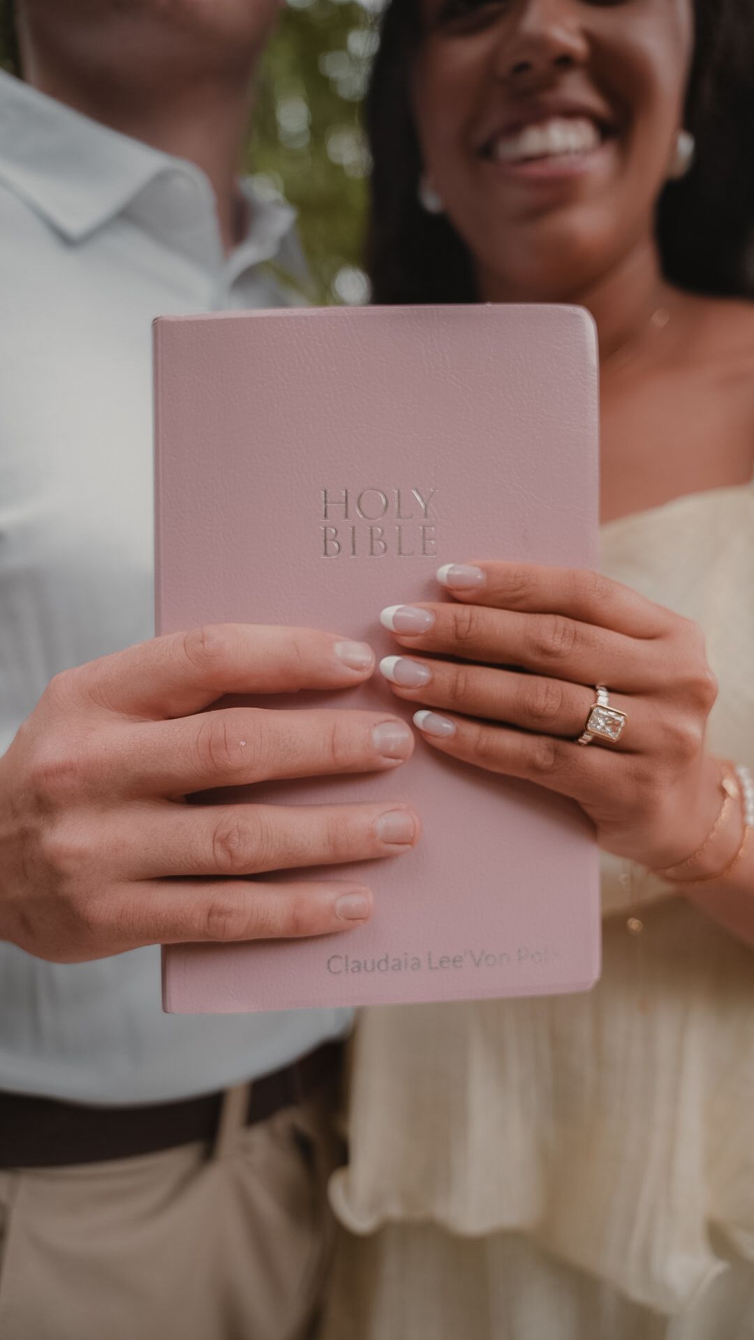 Couple holding a pink personalized Holy Bible with wedding rings visible, smiling outdoors.