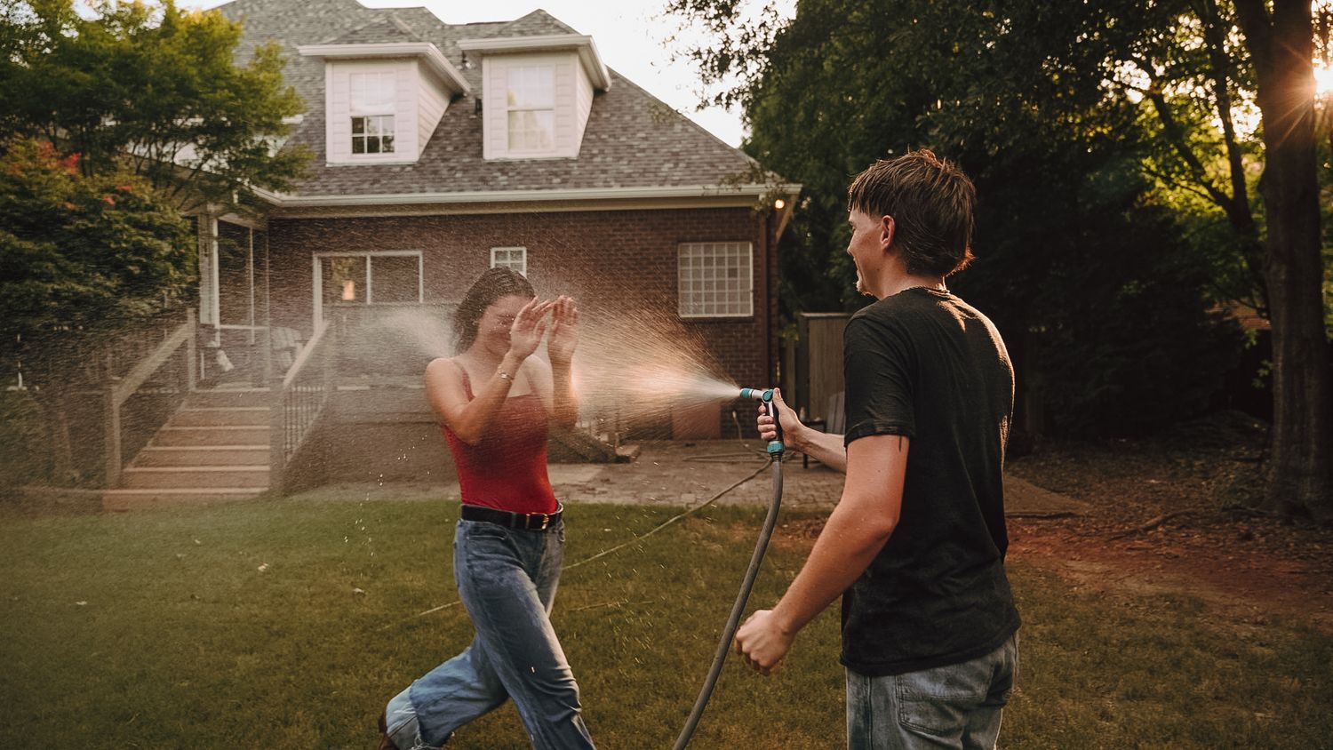 Two young people playing with a garden hose in a sunny backyard, laughing and enjoying summer outdoors.