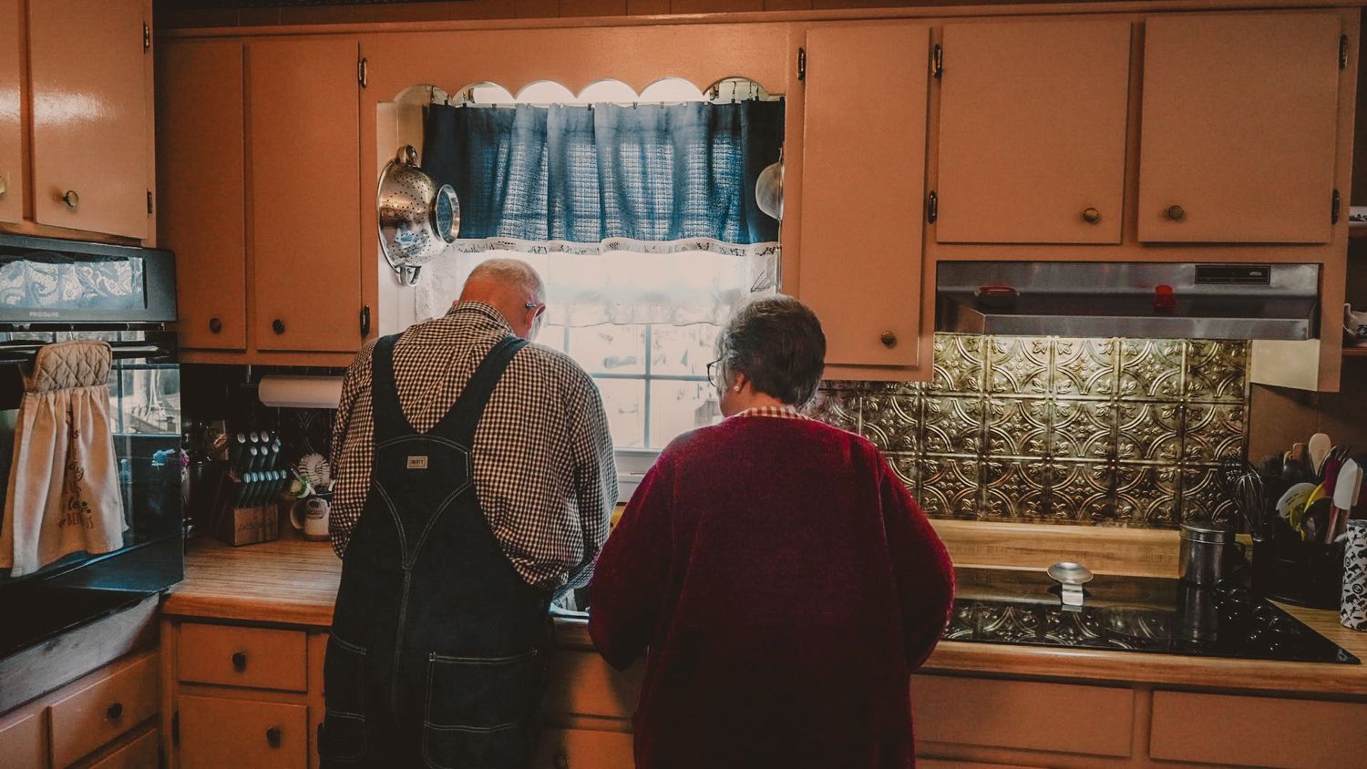 Two elderly people stand together at a vintage kitchen sink with orange cabinets and decorative backsplash.