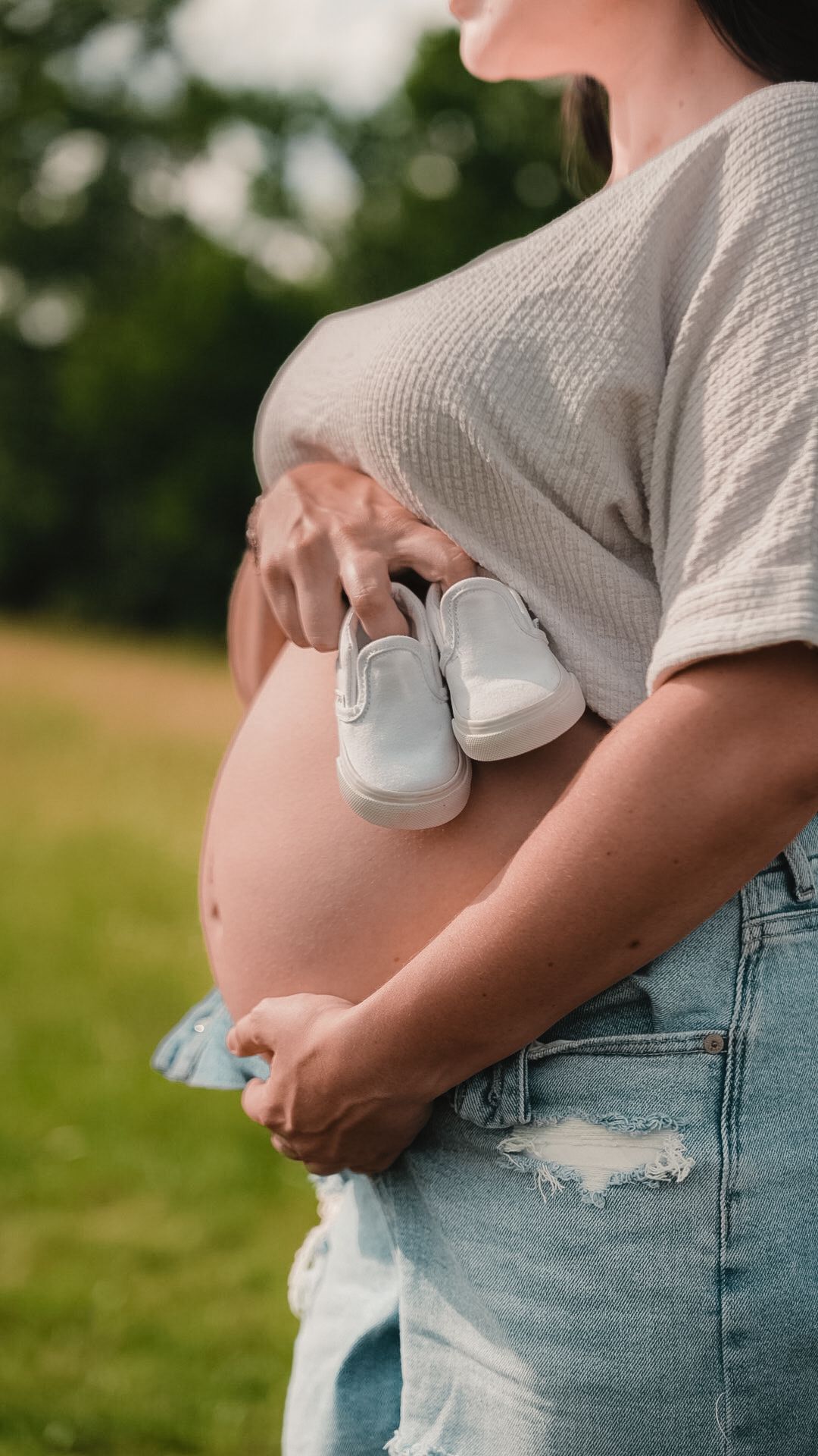 Pregnant woman in ripped jeans holding tiny white baby shoes against her bare bump outdoors.