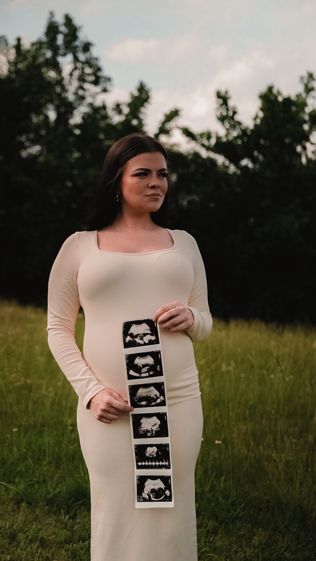 Pregnant woman in white dress holding ultrasound strip outdoors in green field.