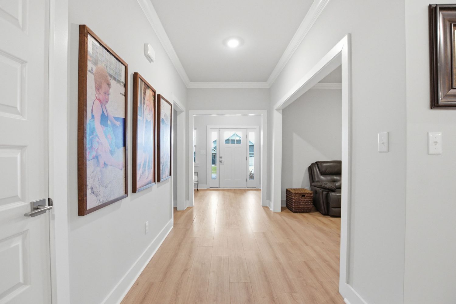 Bright hallway with hardwood floors, white walls, framed artwork, and a glass front door leading to a spacious entryway.