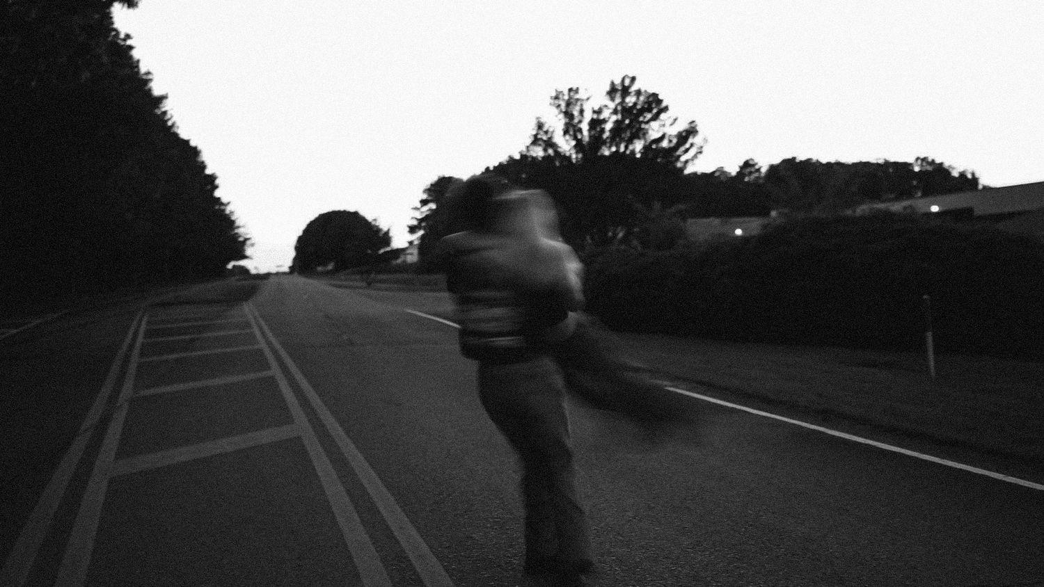 Blurred figure skateboarding on a quiet road at dusk, surrounded by trees, captured in black and white.