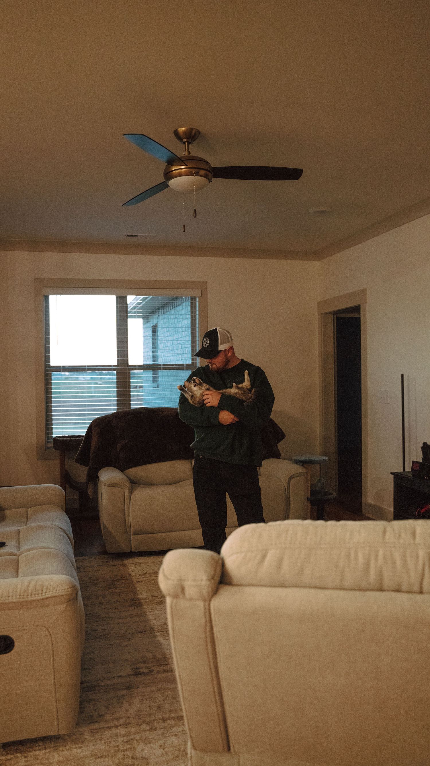Man in baseball cap cradling a cat in a furnished living room with ceiling fan.