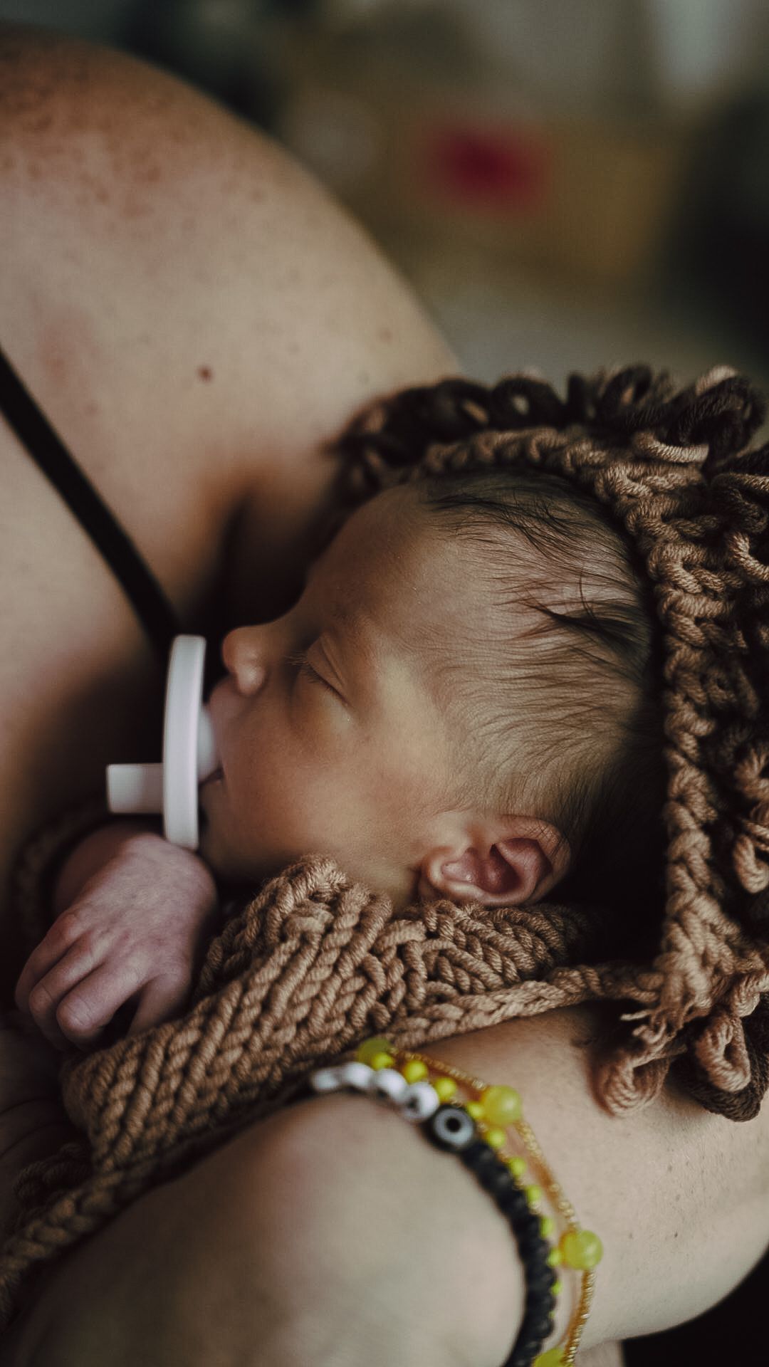 Newborn baby sleeping peacefully on mother's chest, wrapped in knit blanket with pacifier, mother wearing beaded bracelets.