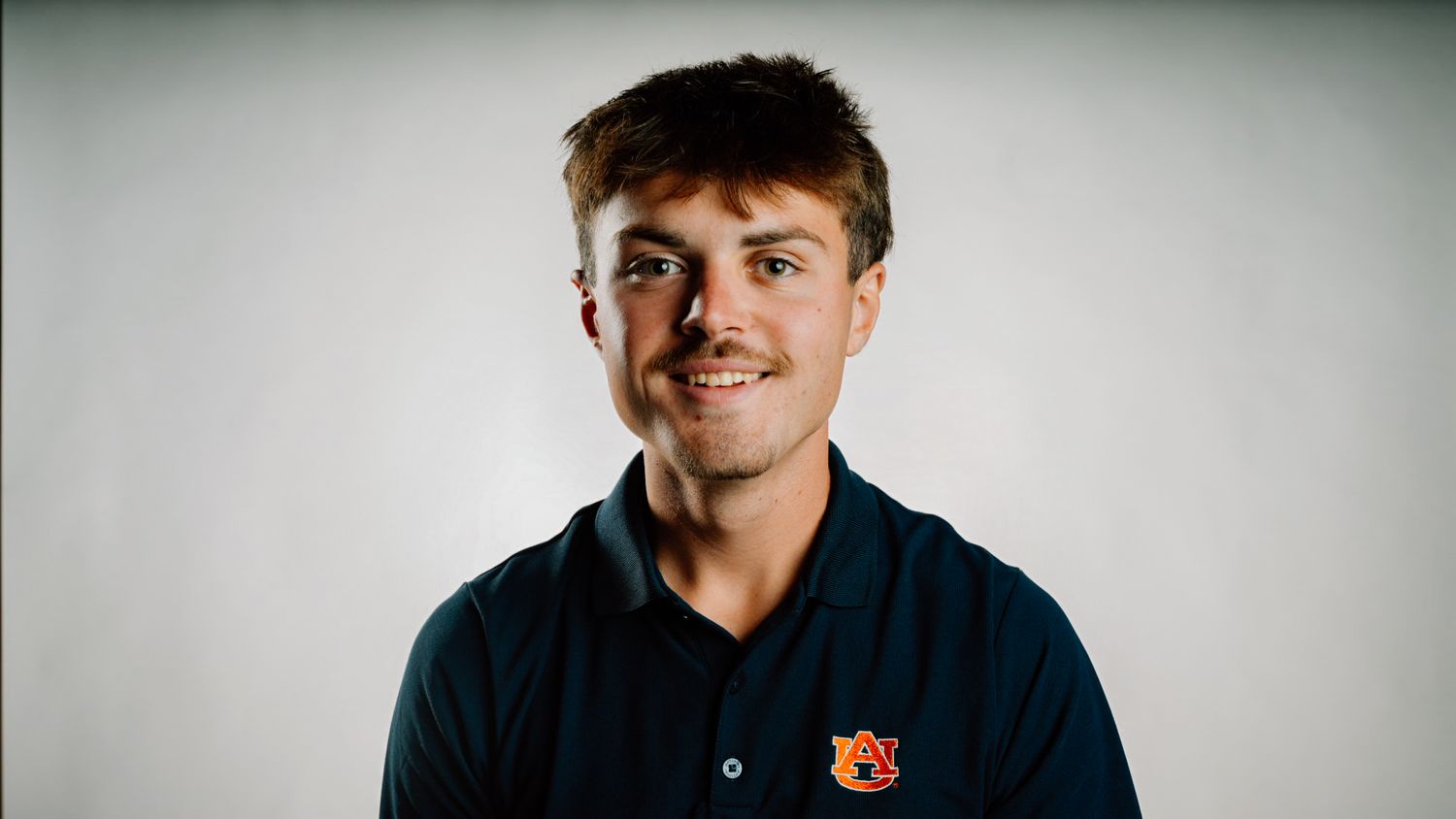 Young man smiling in a navy Auburn University polo shirt against a light gray background.