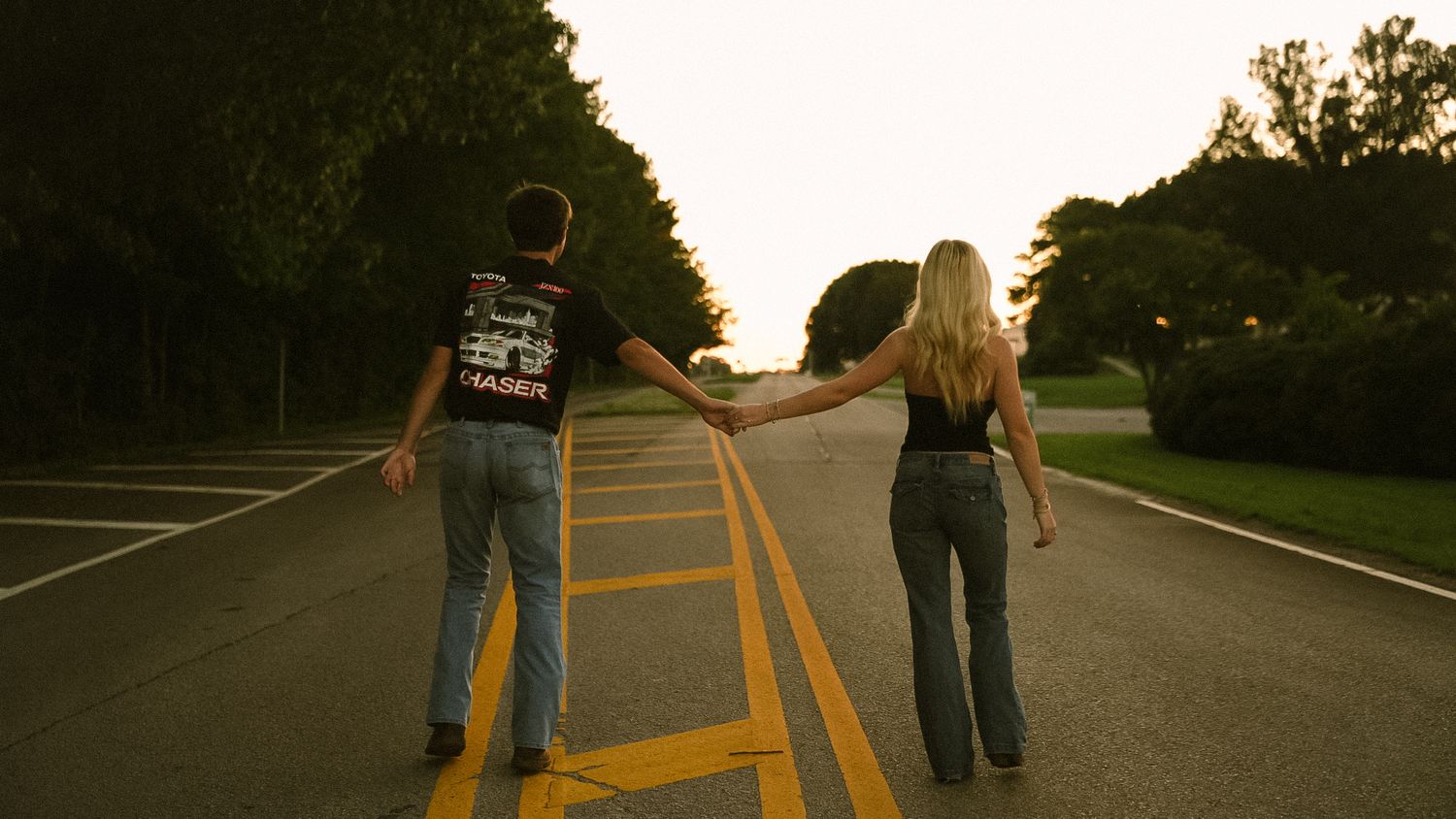 Couple holding hands walking down a tree-lined road at sunset, viewed from behind.