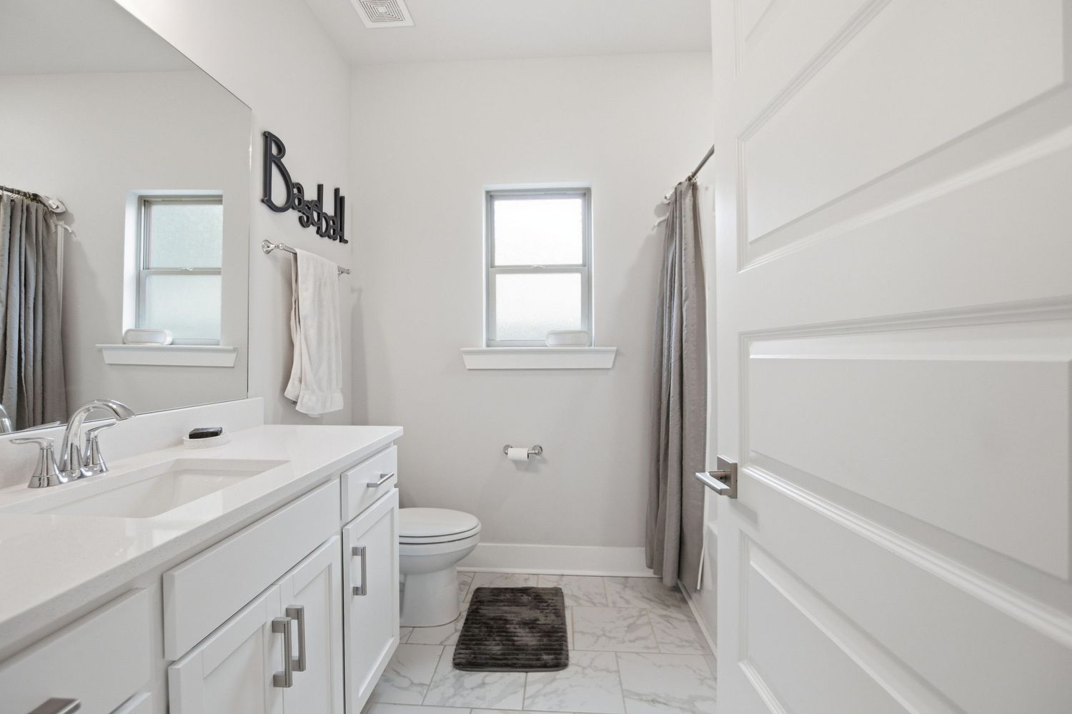 Clean white bathroom with double window, vanity, toilet, and shower with gray curtain.