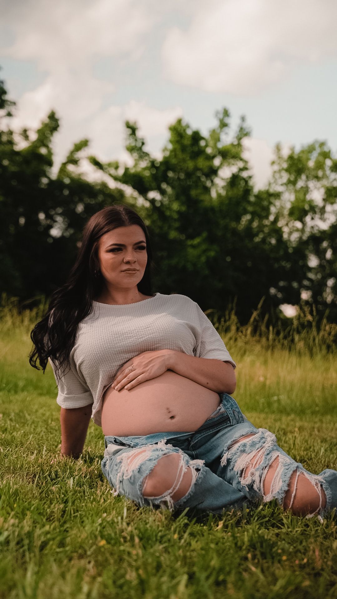Pregnant woman with dark hair sits on green grass cradling bump in white top and ripped jeans.