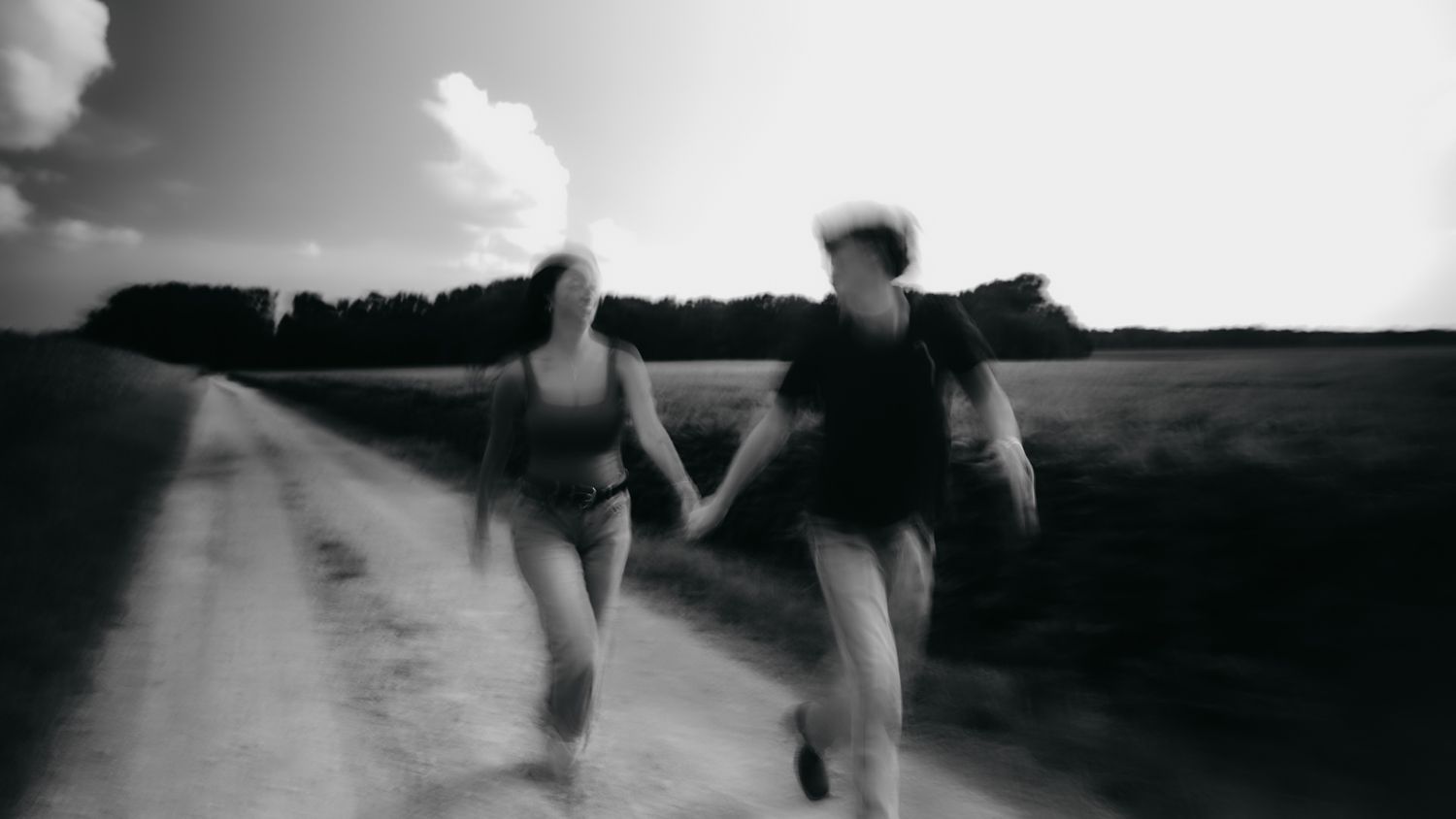 Black and white photo of a couple holding hands running down a rural dirt road surrounded by fields.