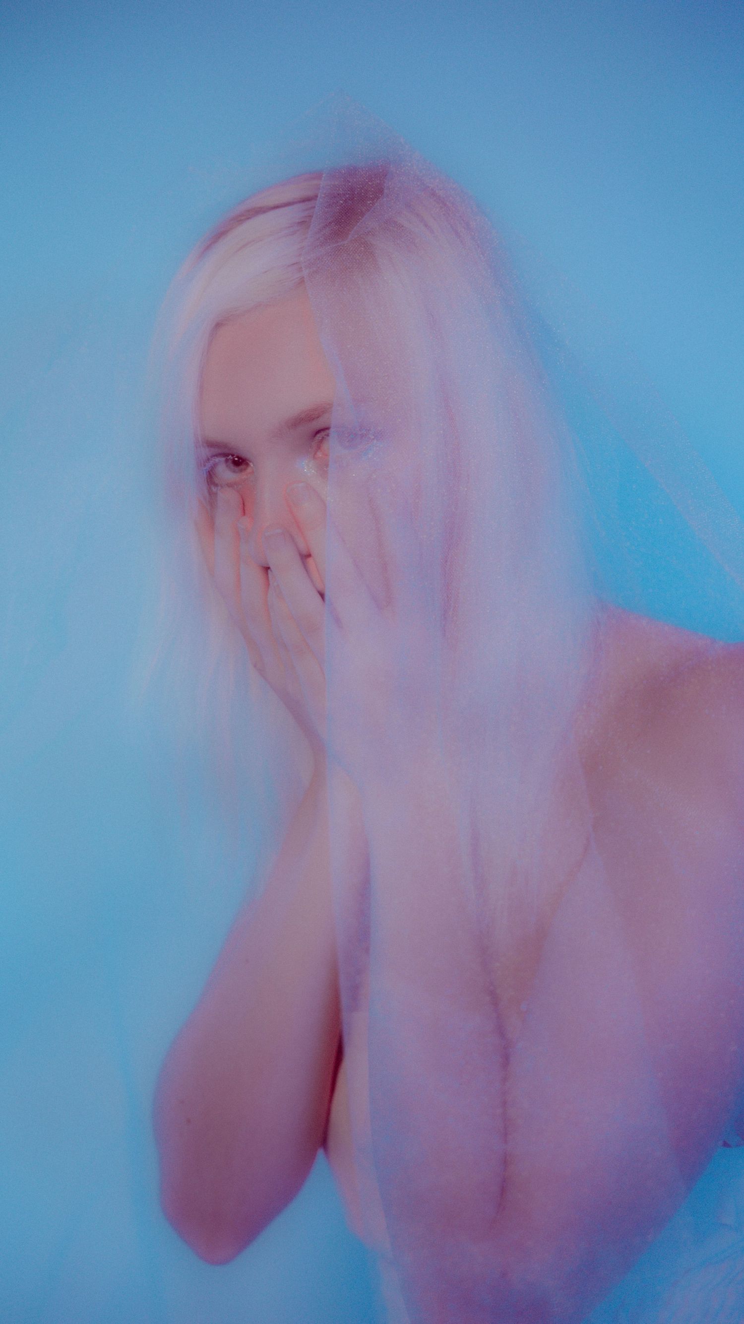 A blond woman with pale skin covers her face with her hands against a soft blue background in an ethereal, dreamy portrait.