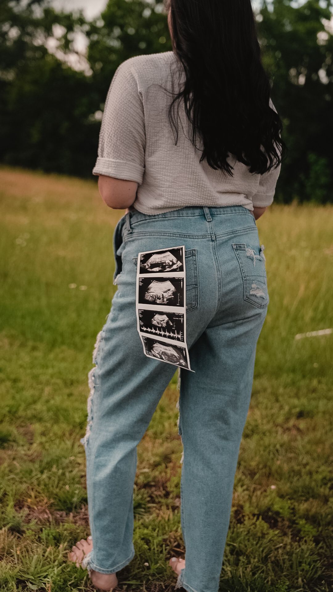 Pregnant woman standing in a field showing ultrasound photos tucked in back pocket of ripped light blue jeans.