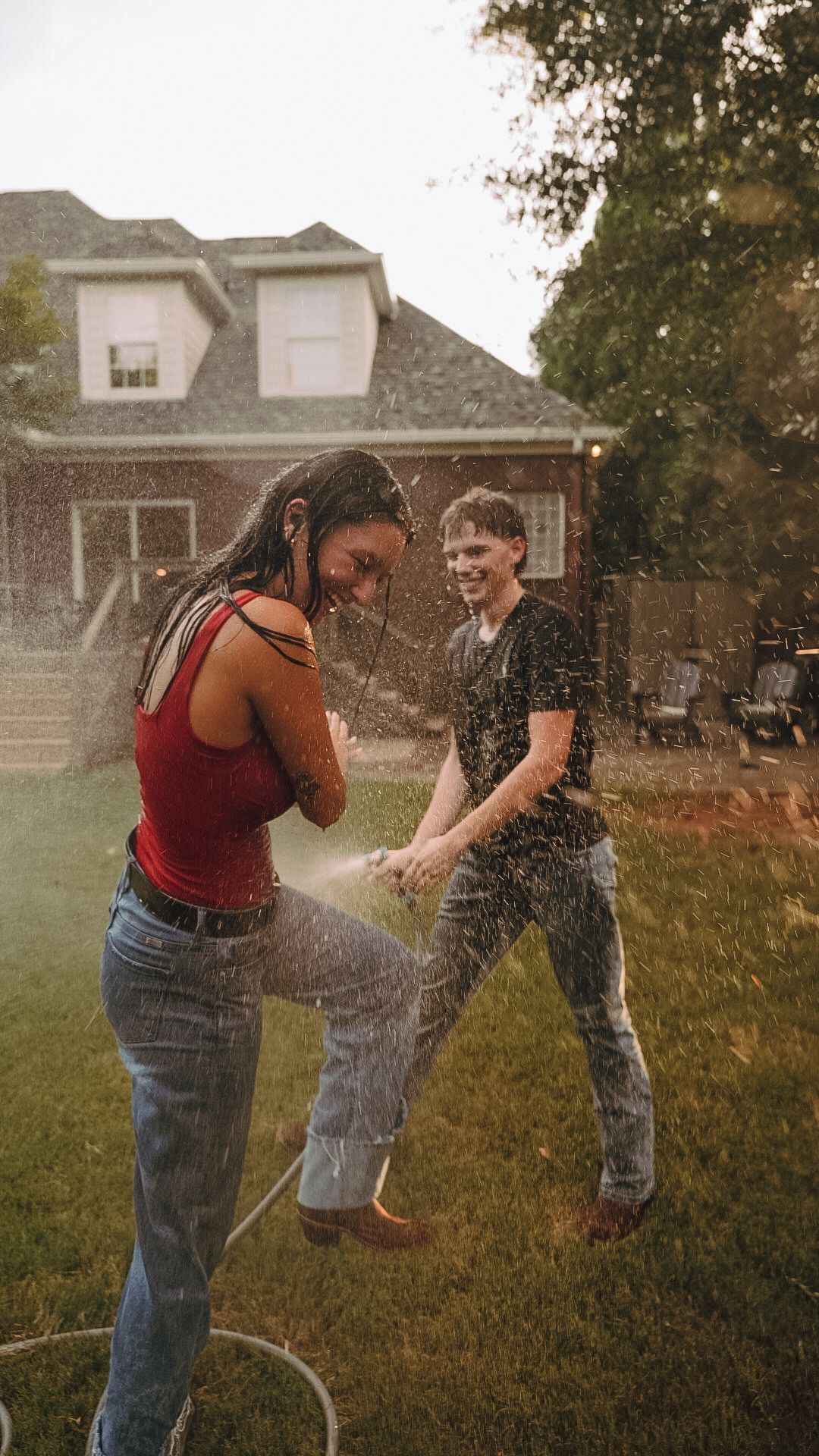 Two young people laughing and playing with a garden hose in a backyard on a warm summer day.