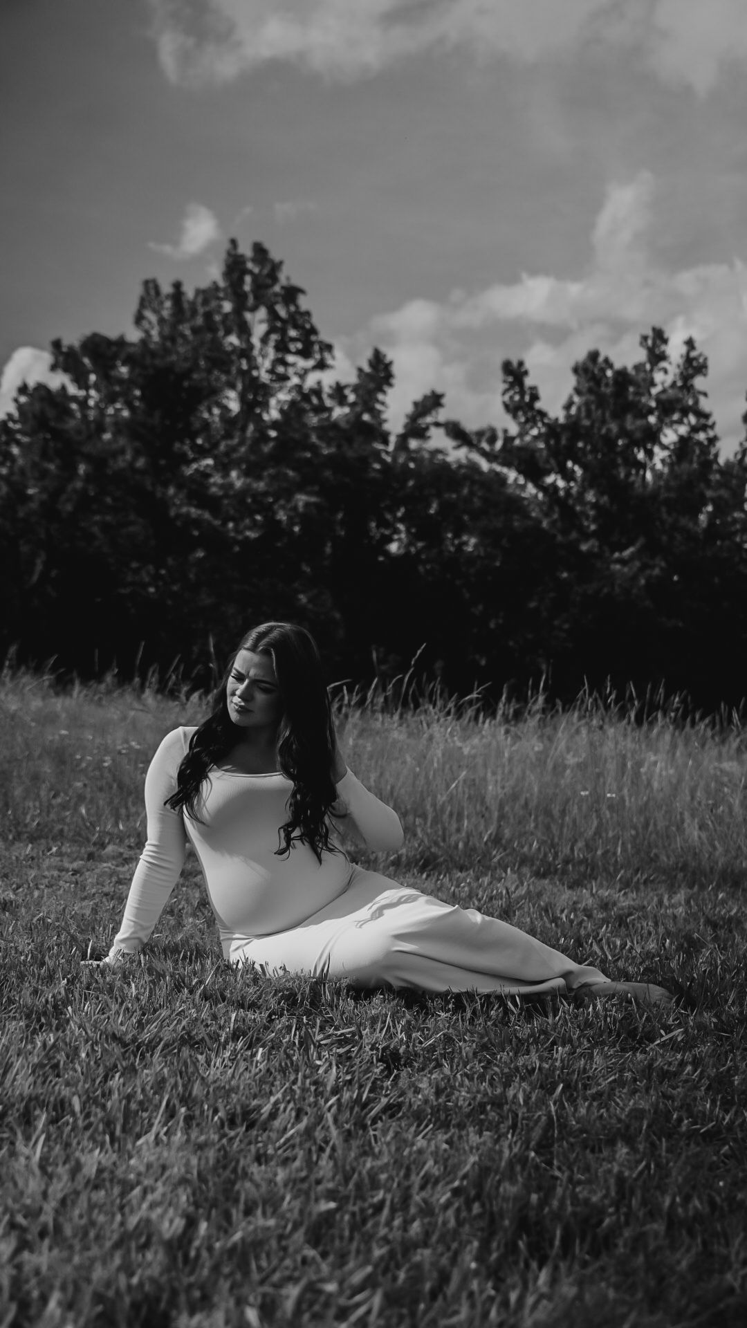 Black and white maternity photo of pregnant woman in white dress sitting in a grassy field.