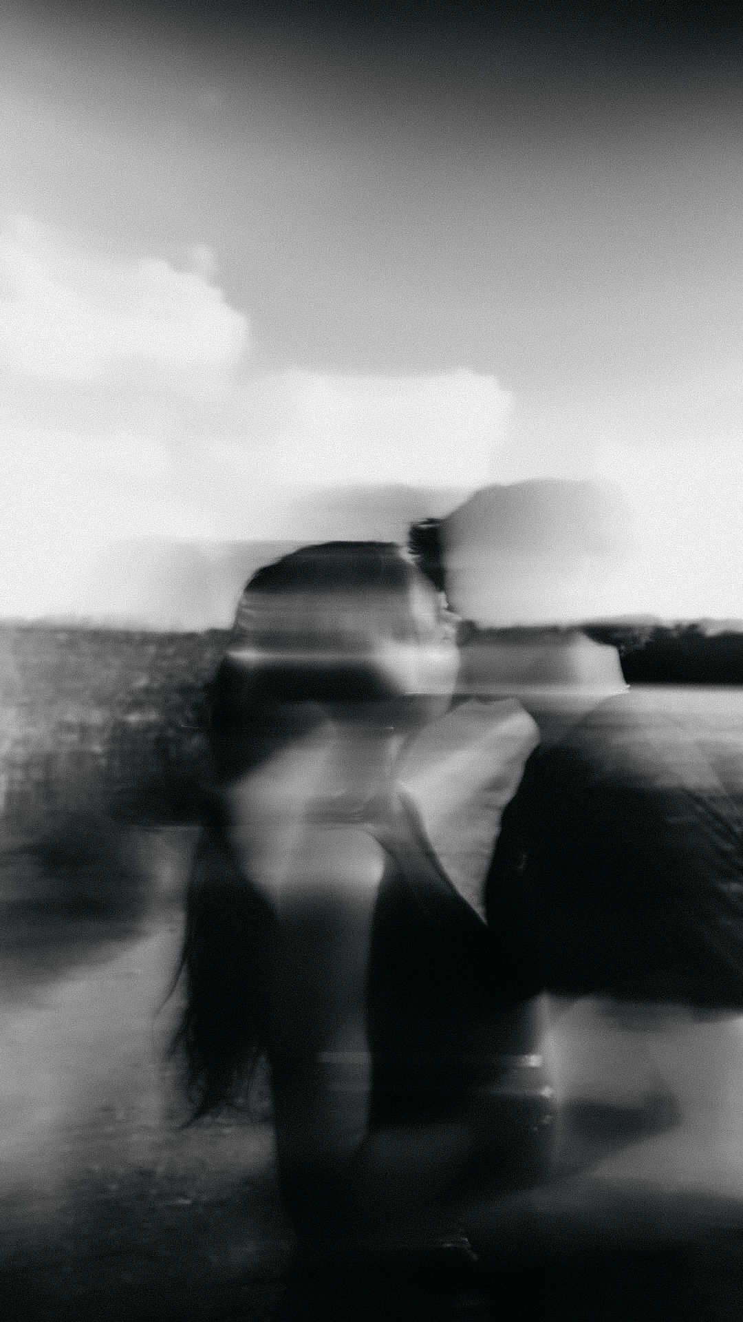 Blurry black and white photo of a person with motion blur effect outdoors under cloudy sky.