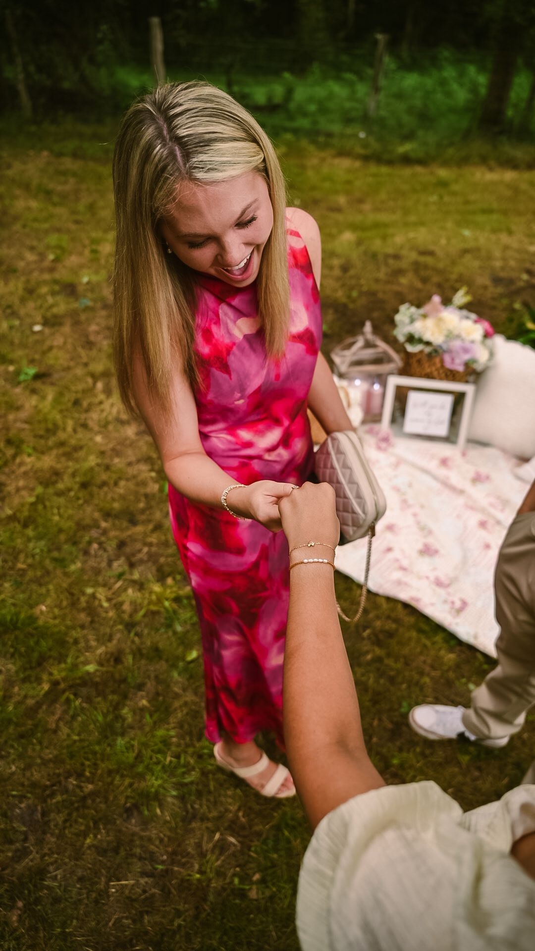 Smiling blonde woman in pink floral dress receiving a bracelet outdoors near a decorated picnic blanket.