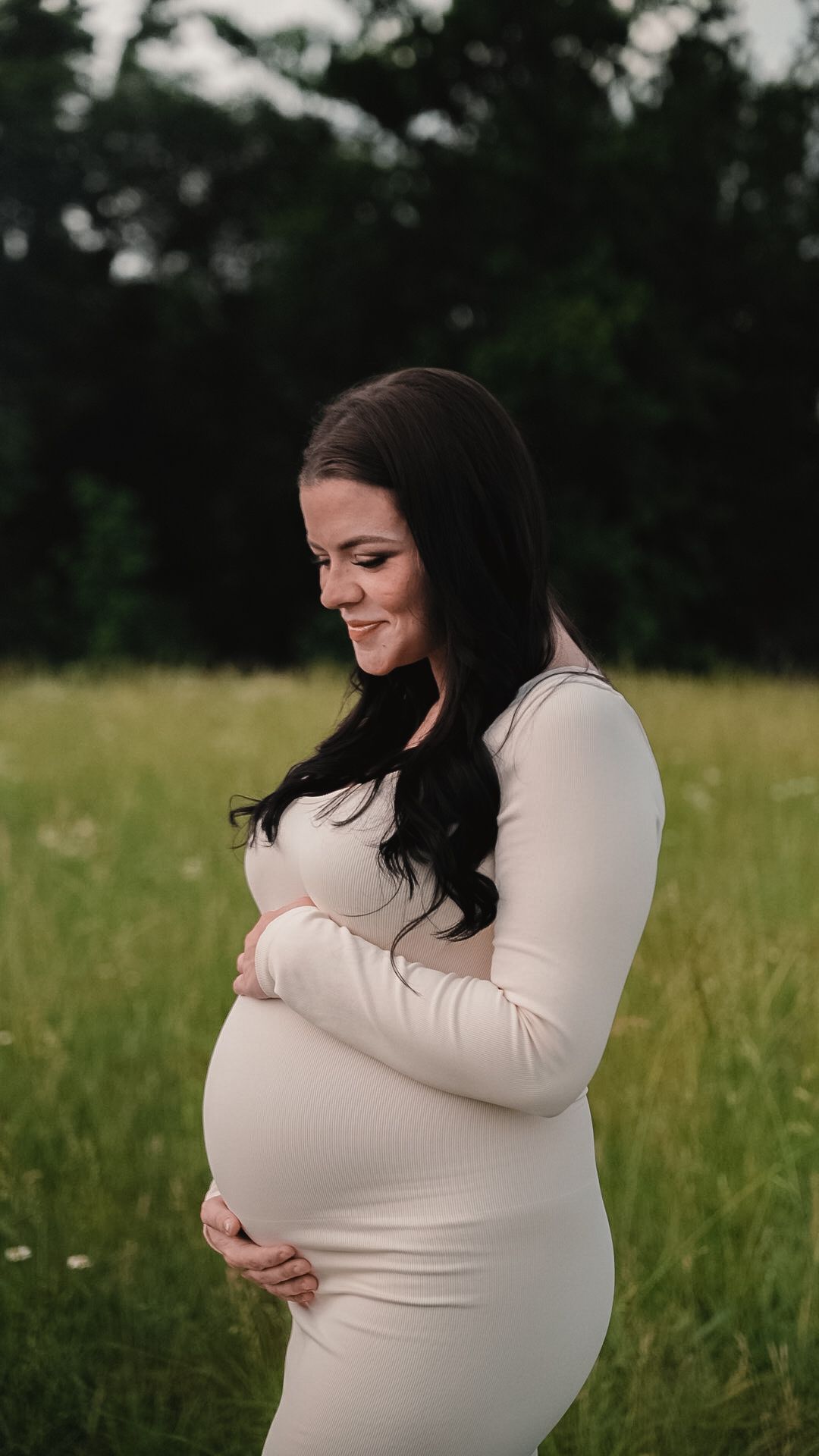 Smiling pregnant woman in white dress cradling baby bump in green field during maternity photo shoot.