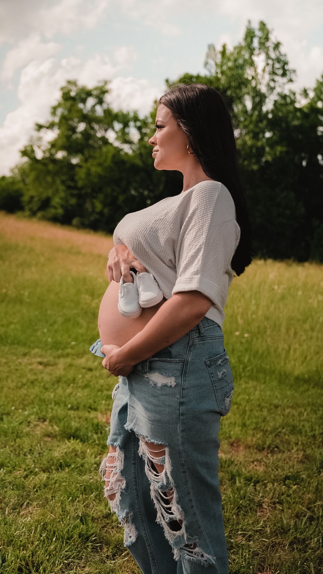 Pregnant woman in ripped jeans holding tiny baby shoes over her bare bump in an outdoor field setting.