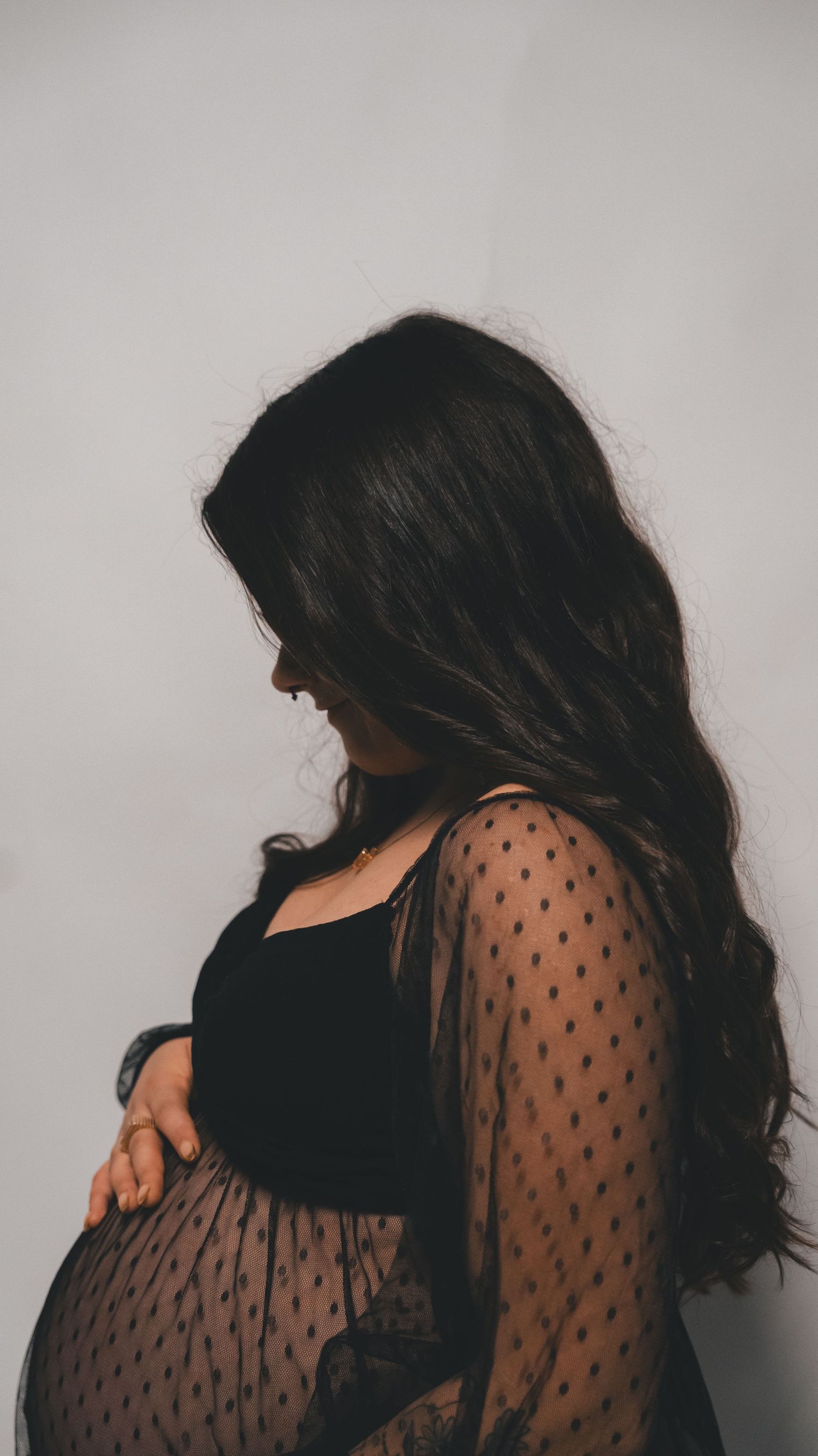 Pregnant woman in black top and sheer polka dot overlay gown cradling her baby bump against a white background.