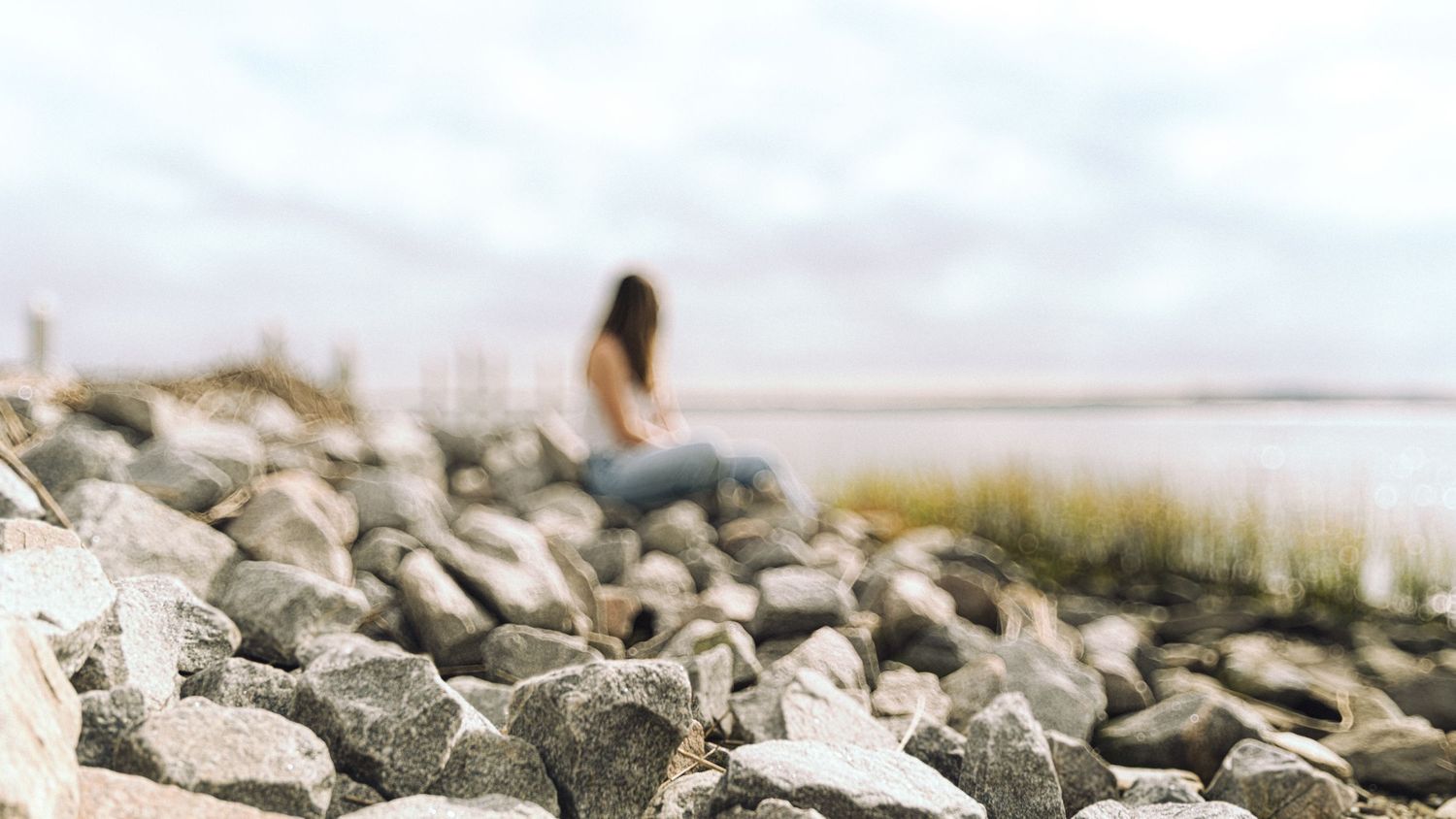 Woman sitting alone on a rocky shoreline with blurred foreground rocks and overcast sky.