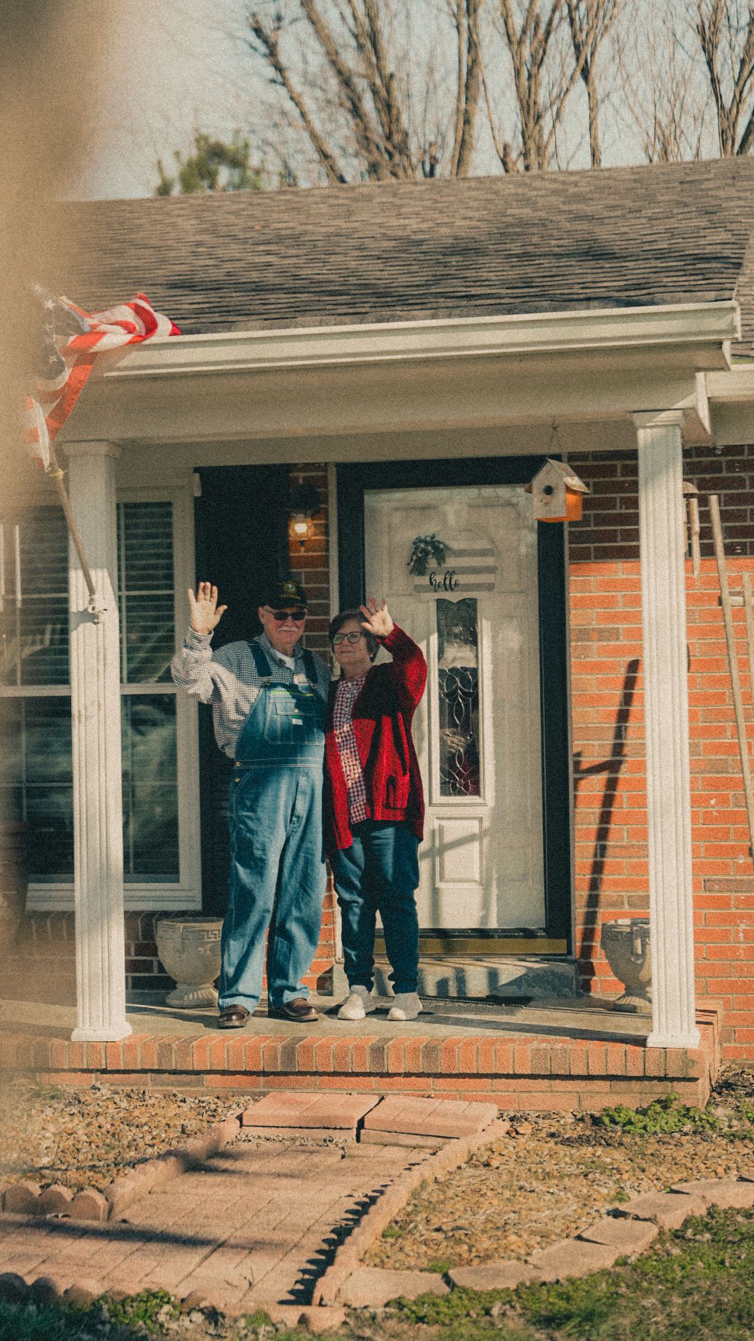 Two people waving from the porch of a brick home decorated with an American flag on a sunny day.