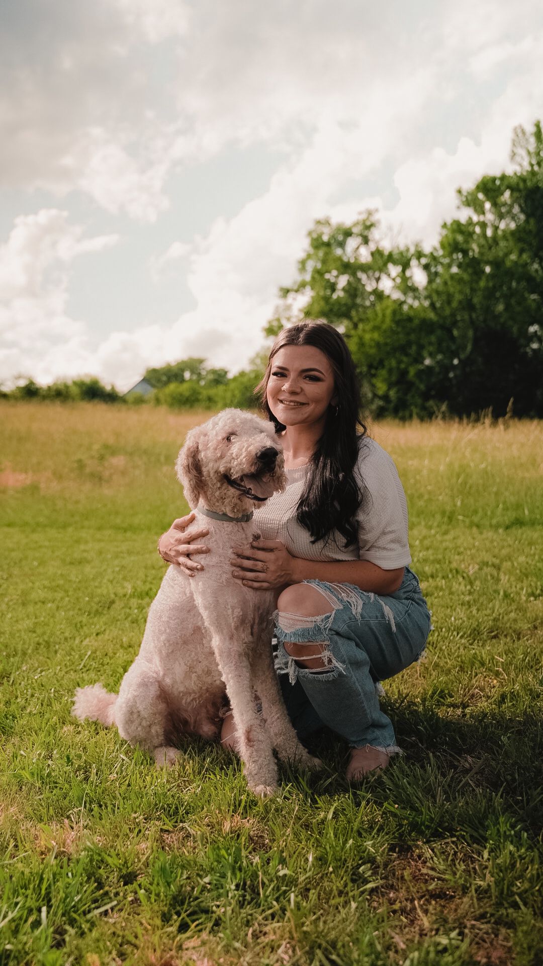 Smiling pregnant woman crouches hugging fluffy goldendoodle dog in lush green outdoor field.