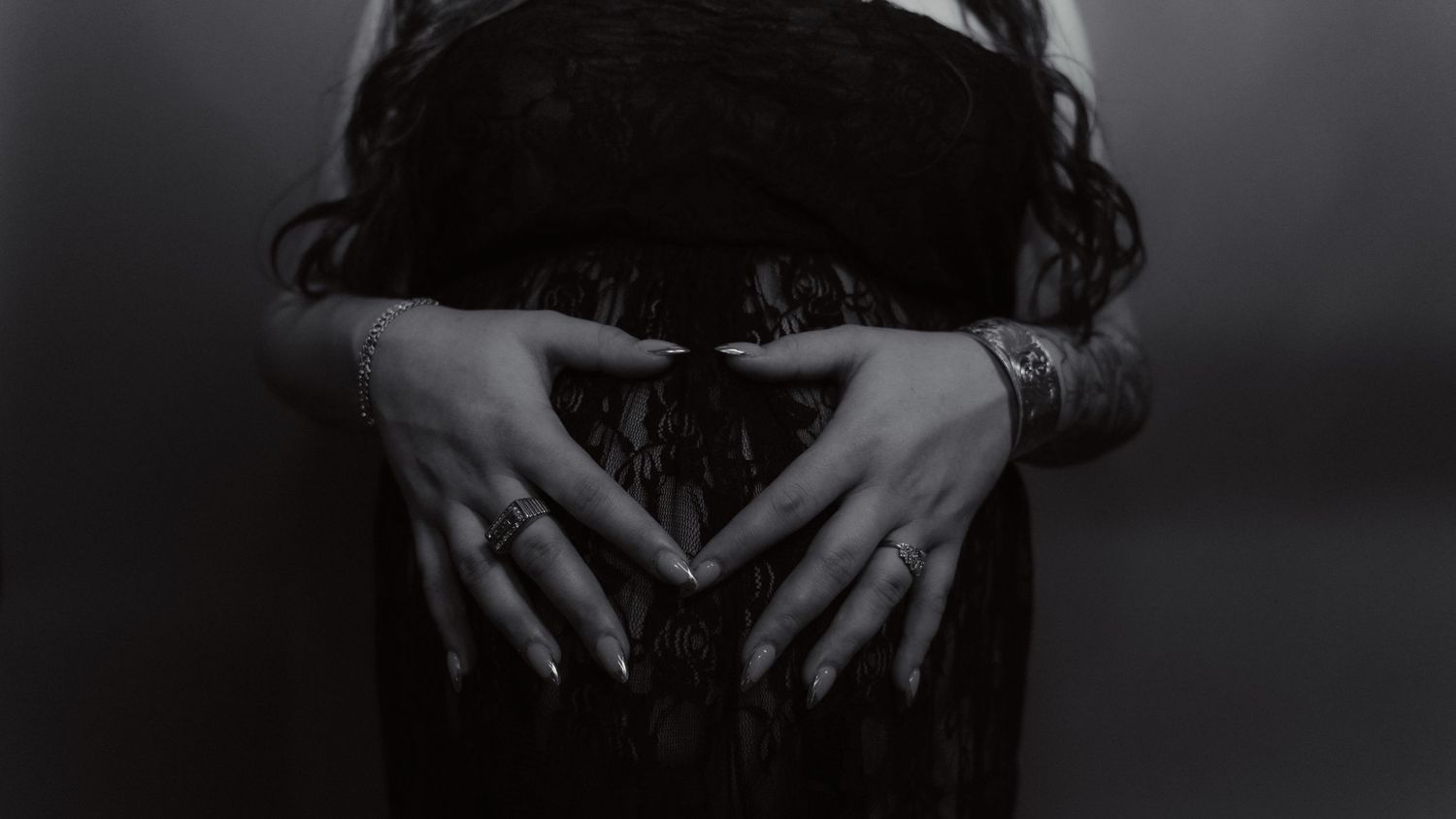 Black and white photo of hands with rings and dark nails forming a heart shape over a dark lace dress.