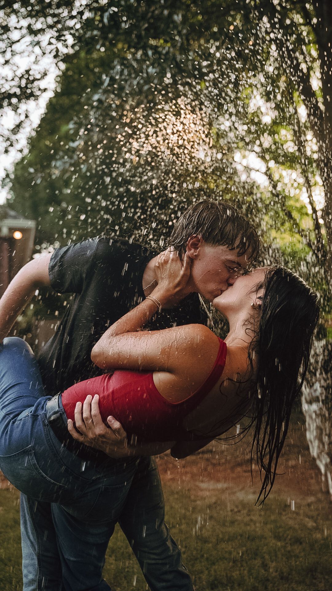 Couple sharing a passionate kiss in the rain, woman in red top being dipped by man in black shirt outdoors.