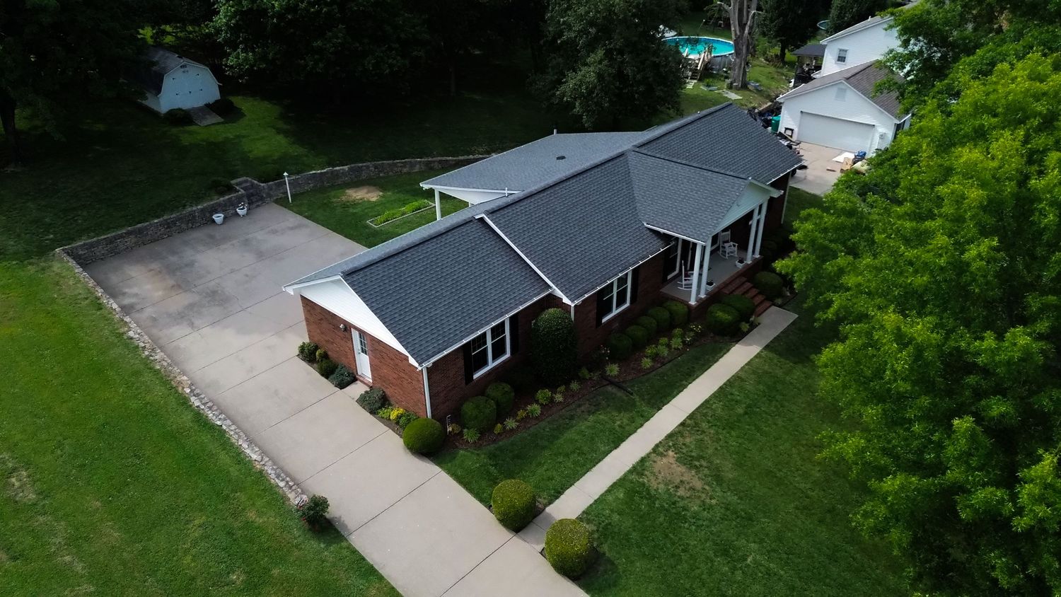Aerial view of a brick ranch-style home with dark roof, circular driveway, and lush green trees surrounding the property.