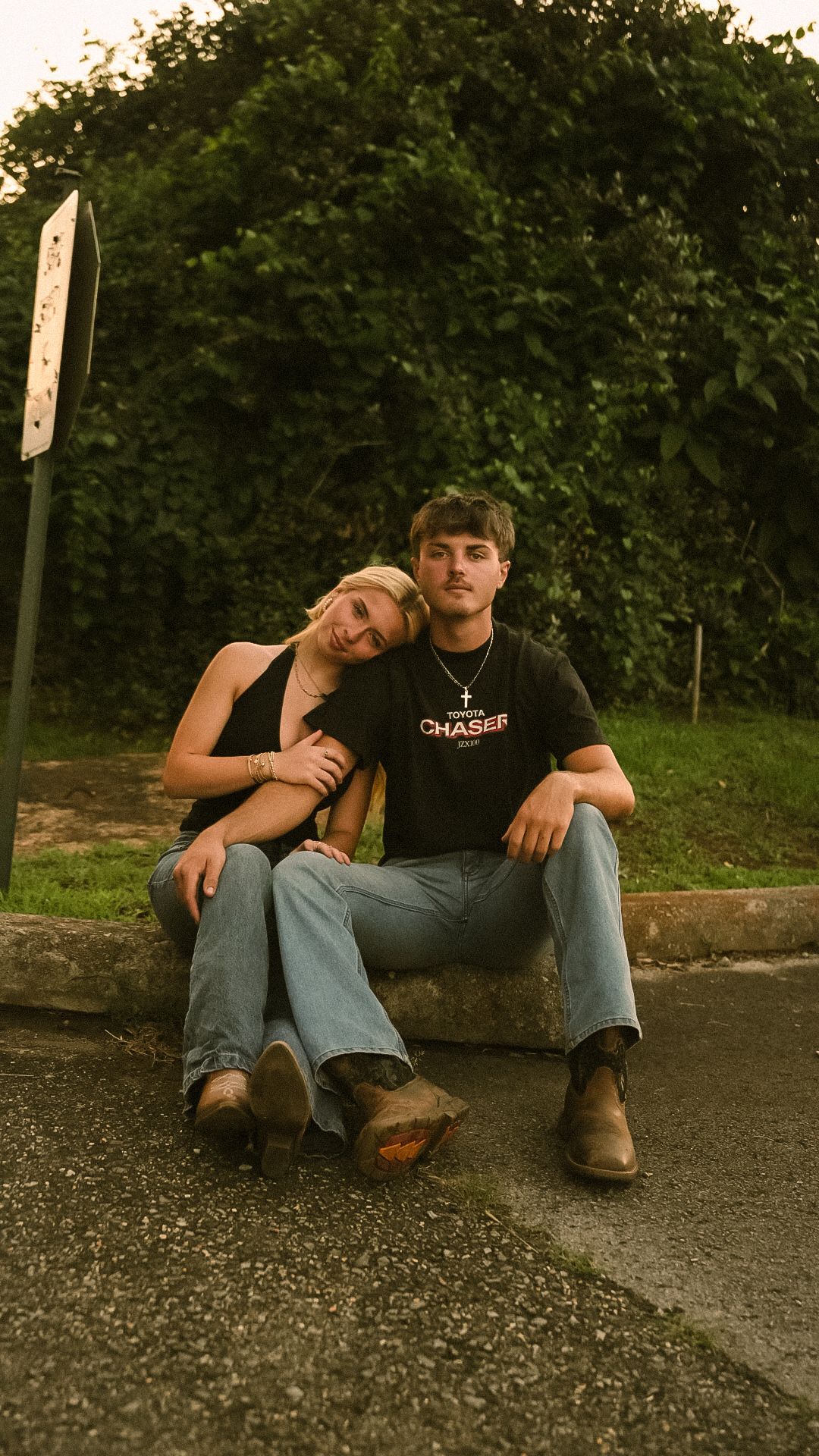 Young couple sitting on a curb outdoors, woman leaning on mans shoulder, both wearing jeans and boots with green trees behind.