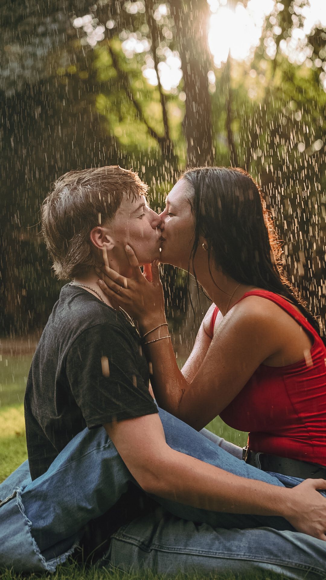 Couple kissing romantically in the rain while sitting on grass in a sunlit park setting.