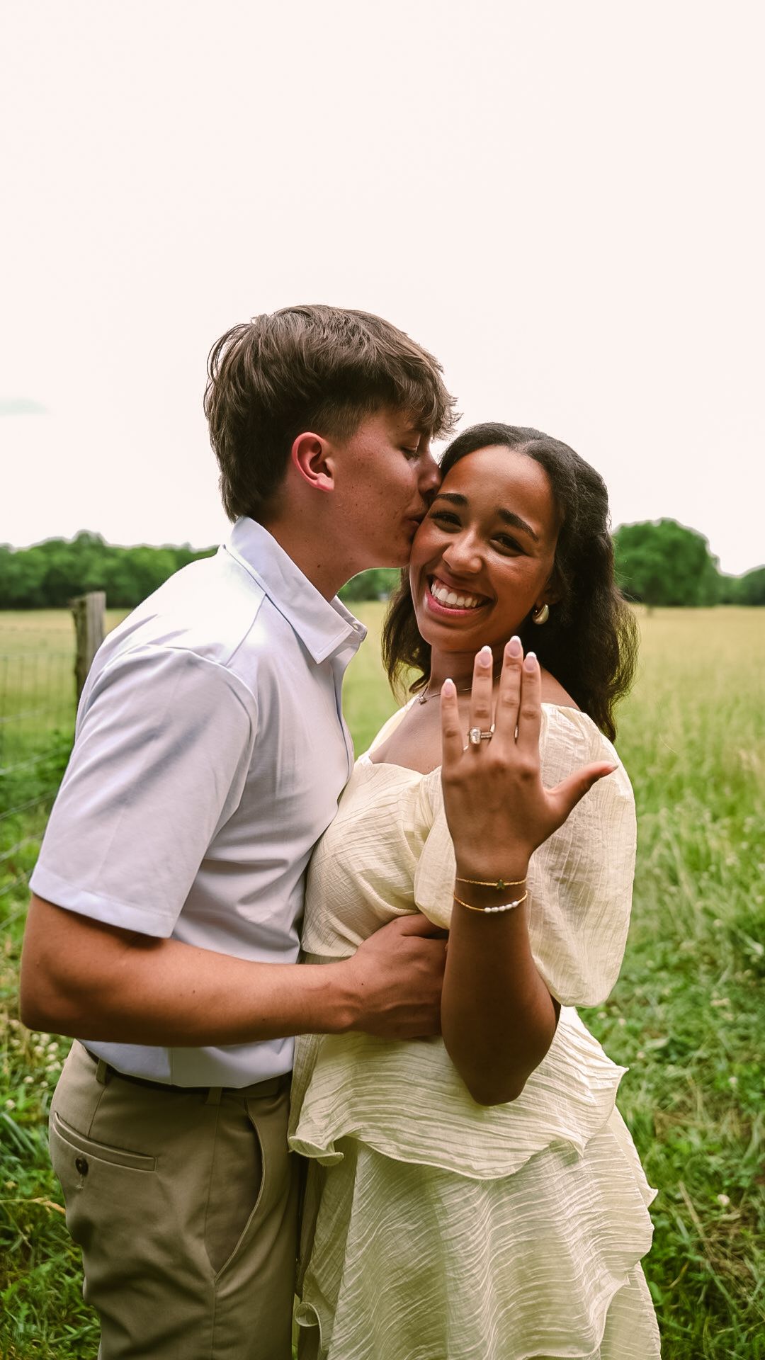 A joyful couple celebrates their engagement in a green field, as the woman shows off her ring while her partner kisses her cheek.