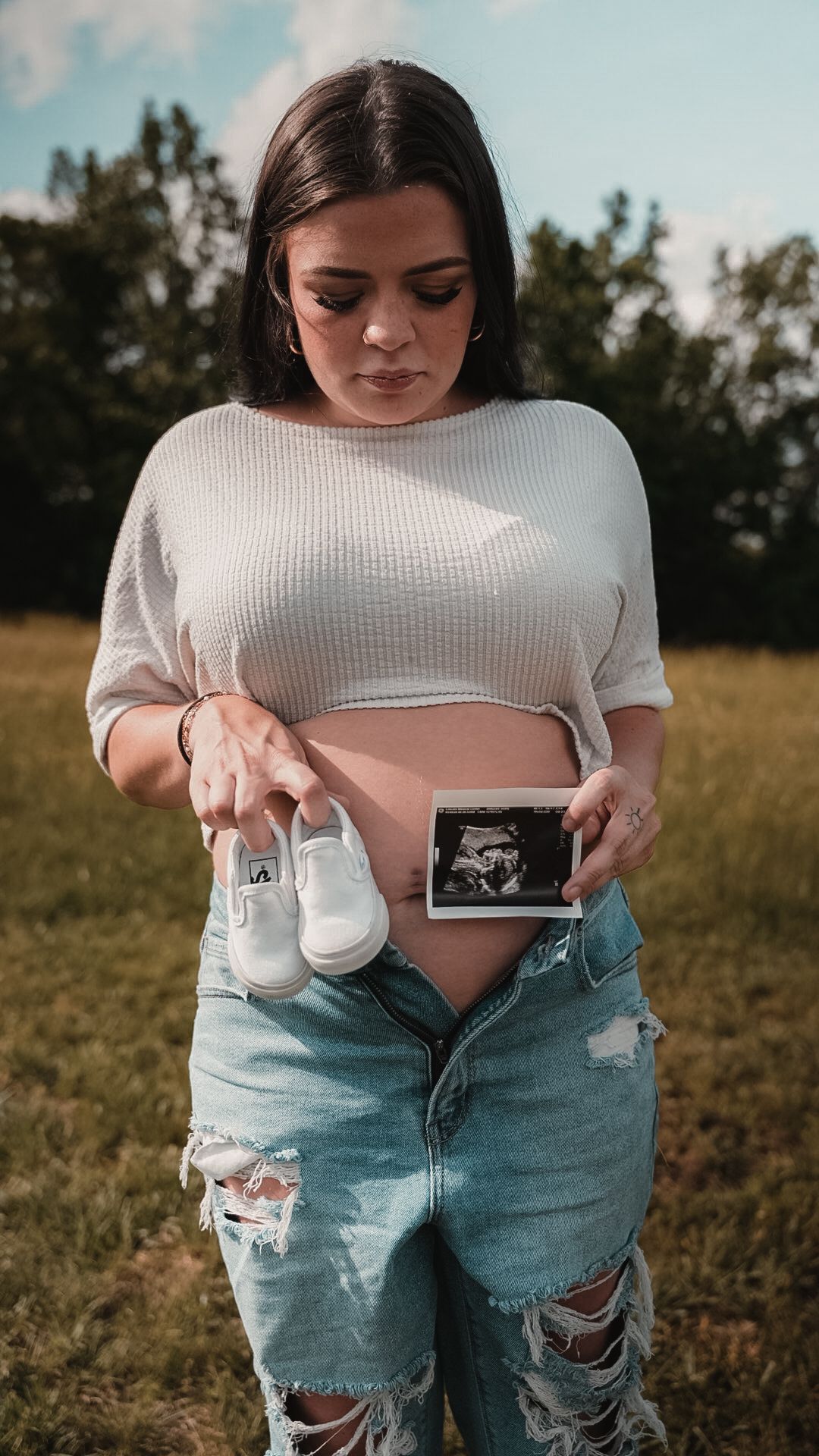 Pregnant woman holding baby shoes and ultrasound photo, wearing crop top and ripped jeans outdoors.